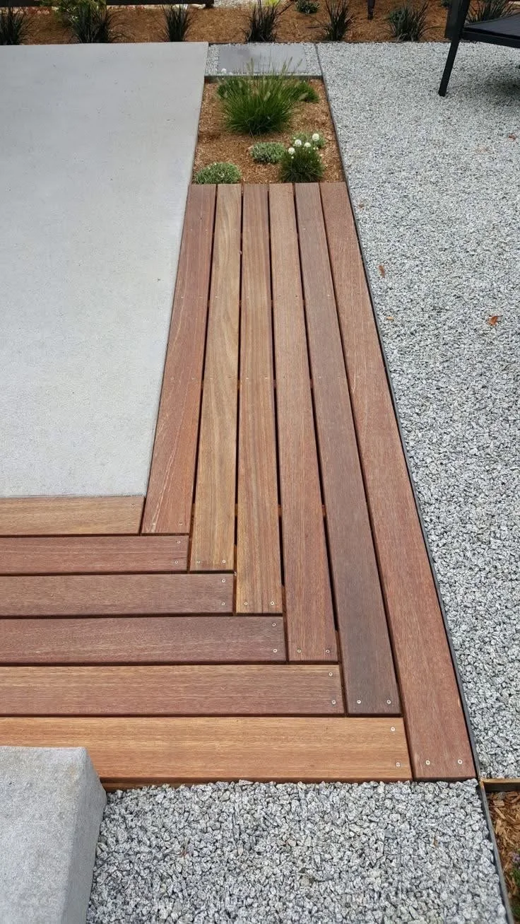Close-up of a modern backyard patio featuring wooden decking, concrete, gravel, and a small garden bed with green plants and white flowers.