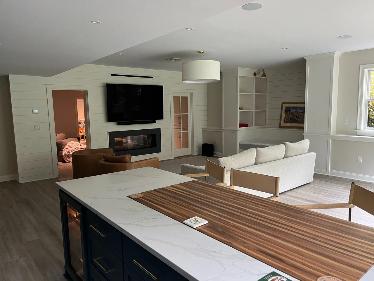 Open-concept living room with white walls, a white sofa, a brown armchair, a wall-mounted flat-screen TV, a fireplace, and large windows with natural light.
