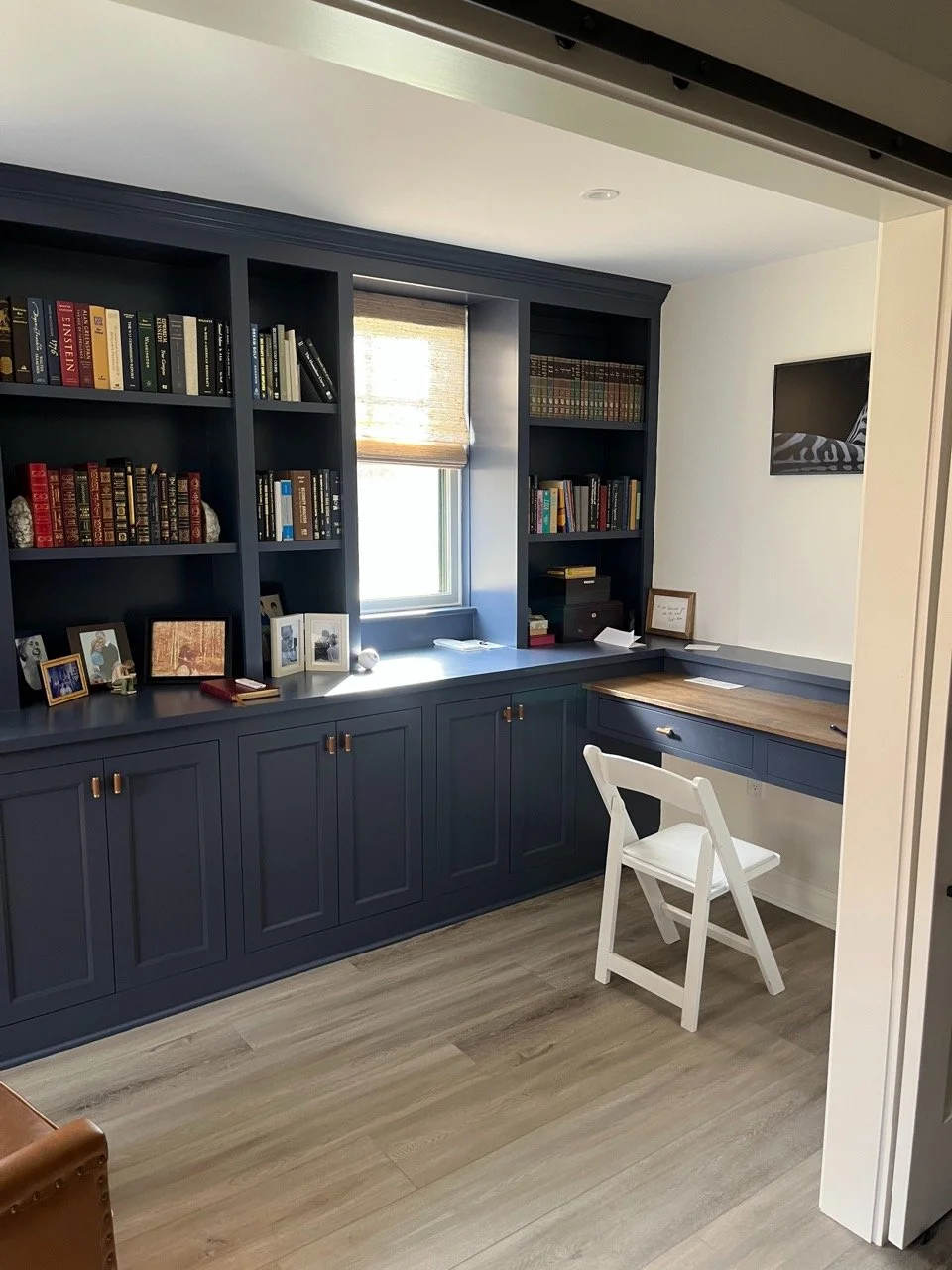 Home library or study room with dark blue built-in bookshelves filled with books. There is a window with a roller shade, a white chair, a wooden desk surface, framed photos, and a television on the wall.