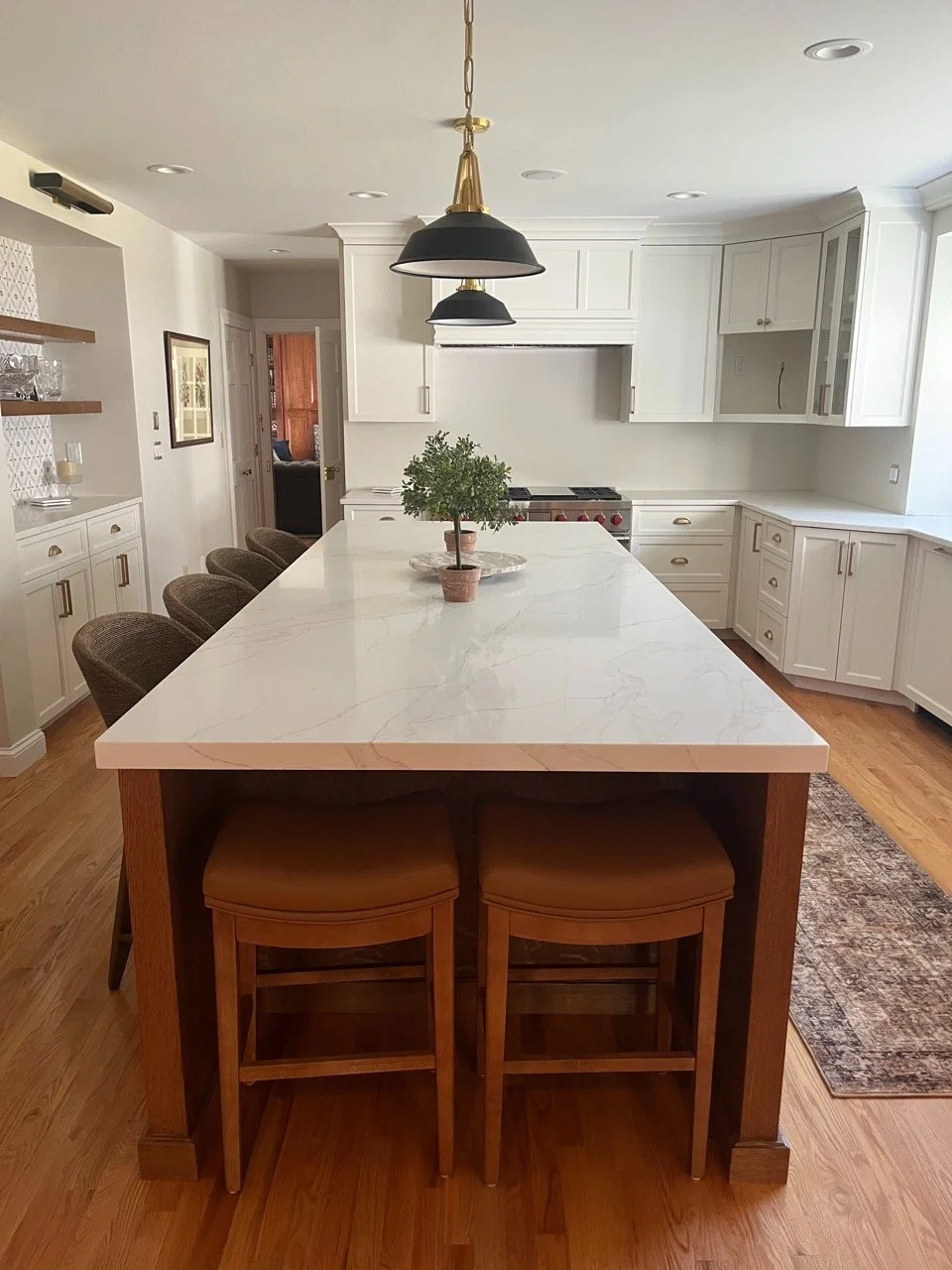 Modern kitchen with a white marble island, two wooden stools, white cabinets, and a plant in the center of the island.