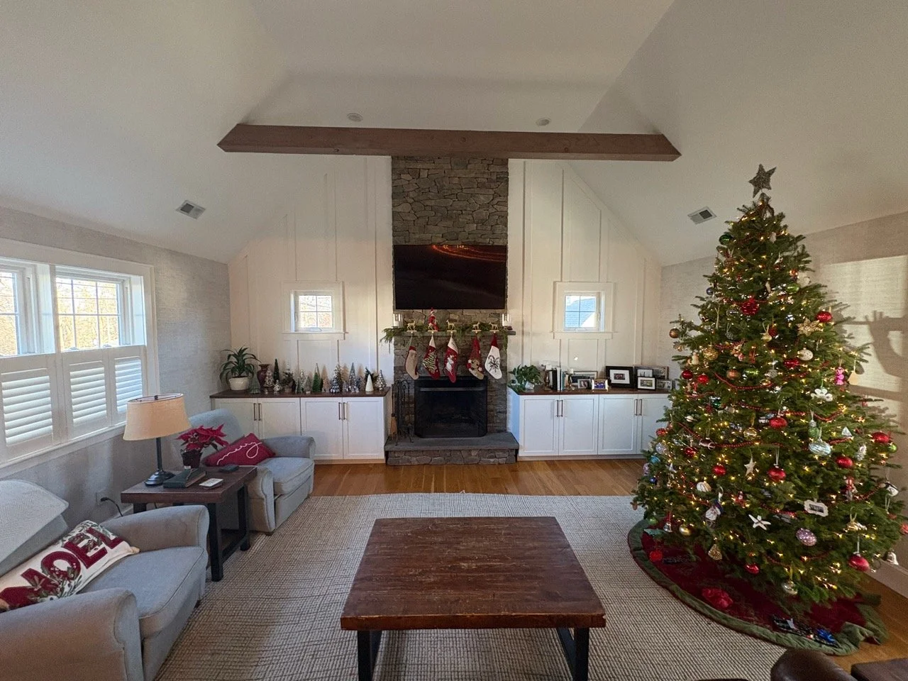 Living room decorated for Christmas with a Christmas tree, stockings hanging above the fireplace, and holiday decorations on the side cabinets.