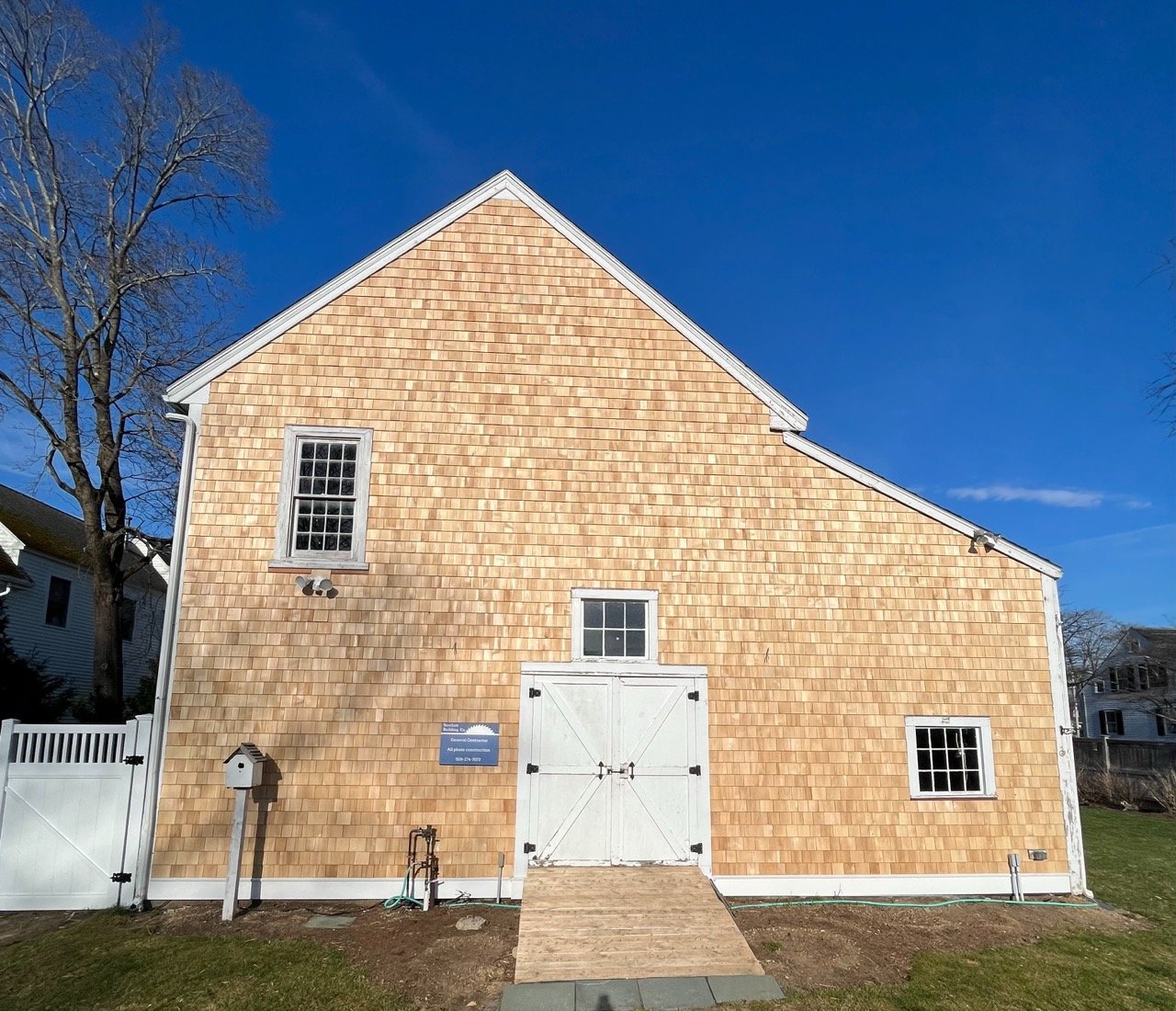 Barn with new light wood siding, white doors and windows, a ramp leading to the door, a small white fence on the left, and a clear blue sky.