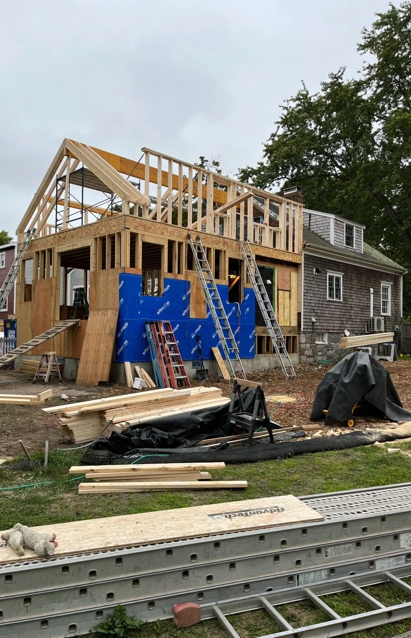Construction site of a two-story house with wooden framing and scaffolding, surrounded by construction materials and equipment.