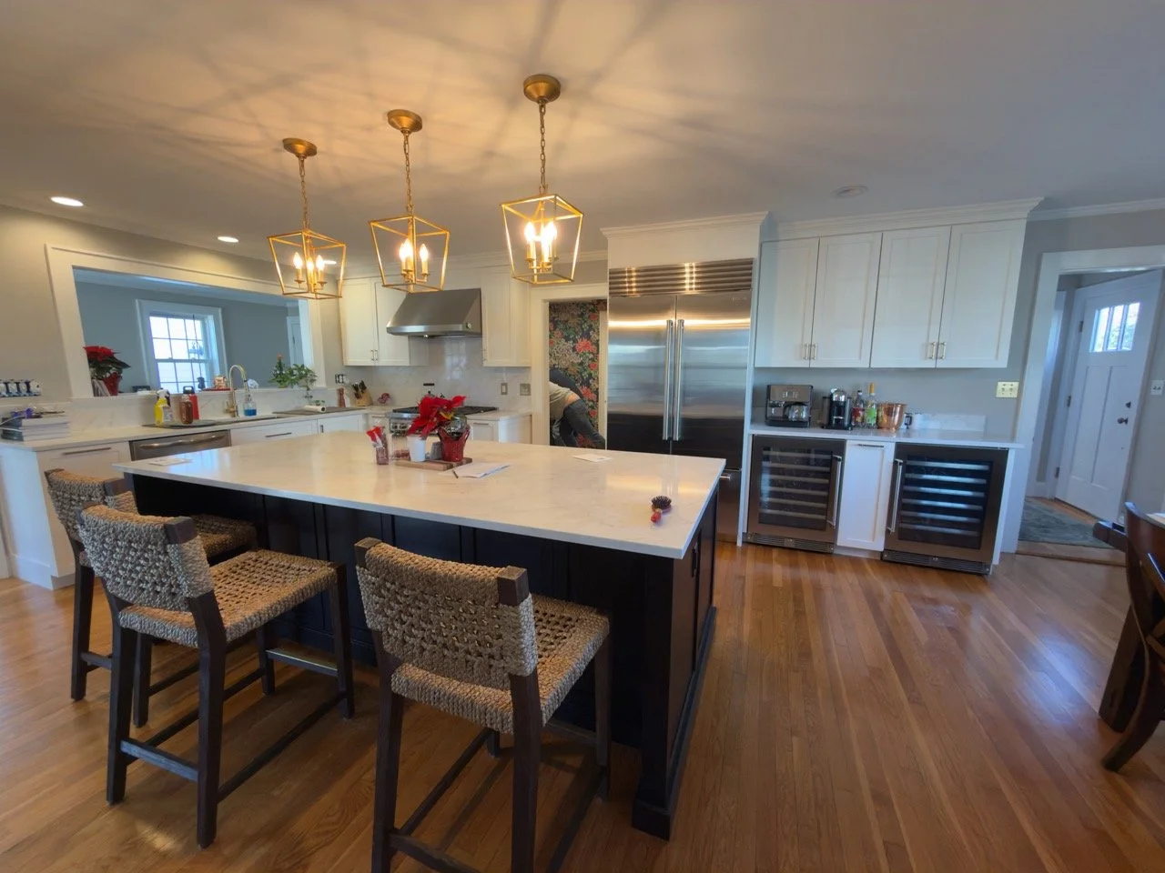 Modern kitchen with white cabinets, stainless steel appliances, and a large island with a marble countertop. Three pendant lights hang above the island, and there are four woven chairs around it. The floor is wooden, and there are holiday decorations