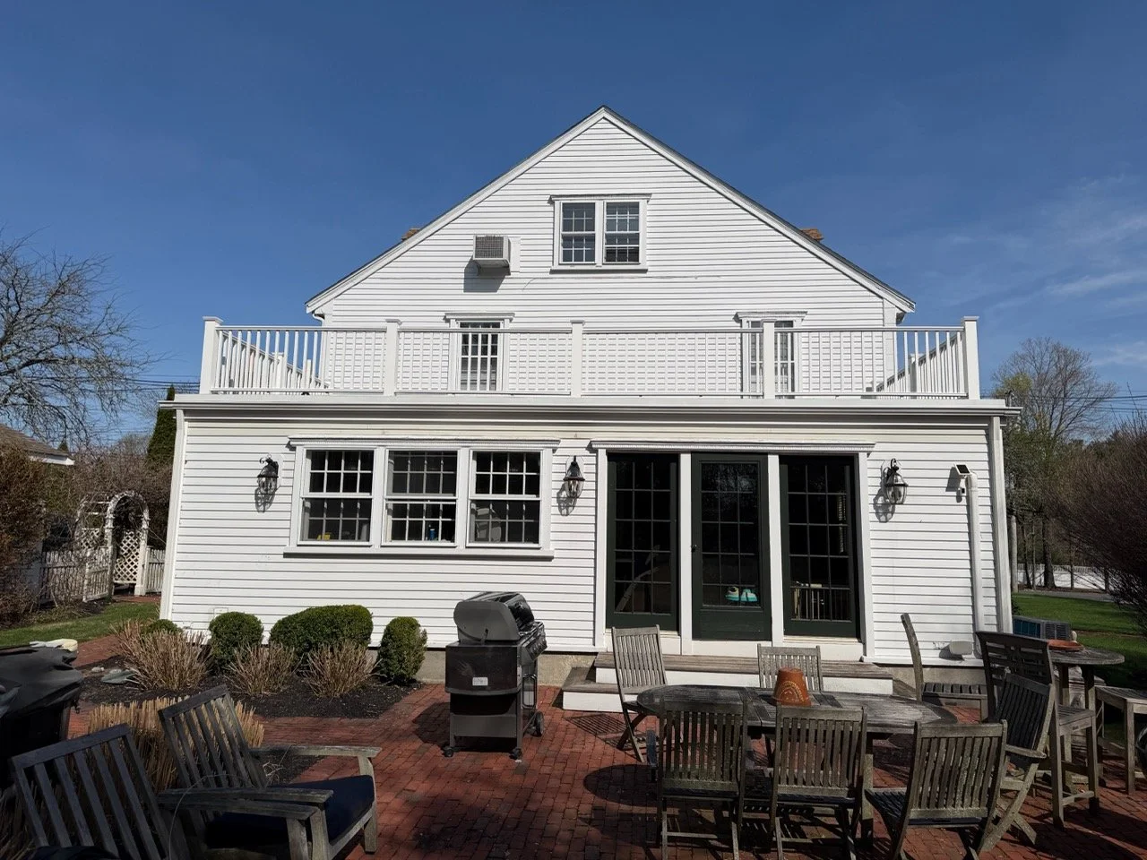 A white two-story house with a balcony, multiple windows, and outdoor patio furniture on a brick patio, with a grill and bushes in the yard, under a clear blue sky.