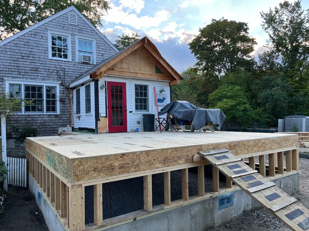 Construction site of a small house with a wooden deck and stairs, surrounded by trees, during daytime.