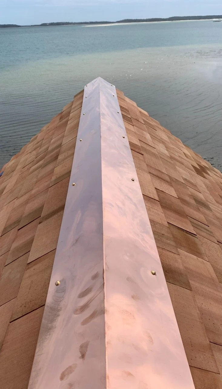Close-up view of a copper ridge cap on a shingled roof beside a large body of water, with a distant shoreline under a cloudy sky.
