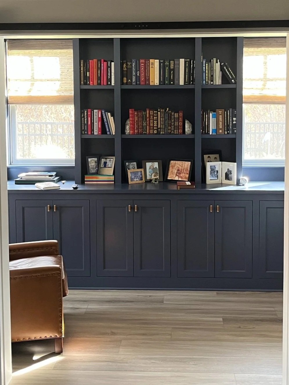 Blue bookshelf with framed photos and books, windows in background, wooden floor, and leather armchair to the left.