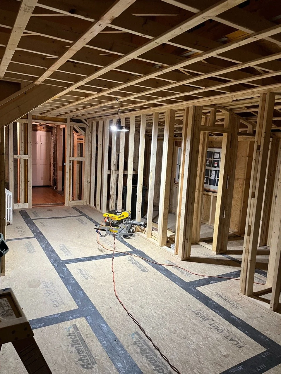 Interior of a house under construction with exposed wooden framing, unfinished walls, and a concrete subfloor with black tape markings. There is a work light hanging from the ceiling and a yellow power saw on the floor.
