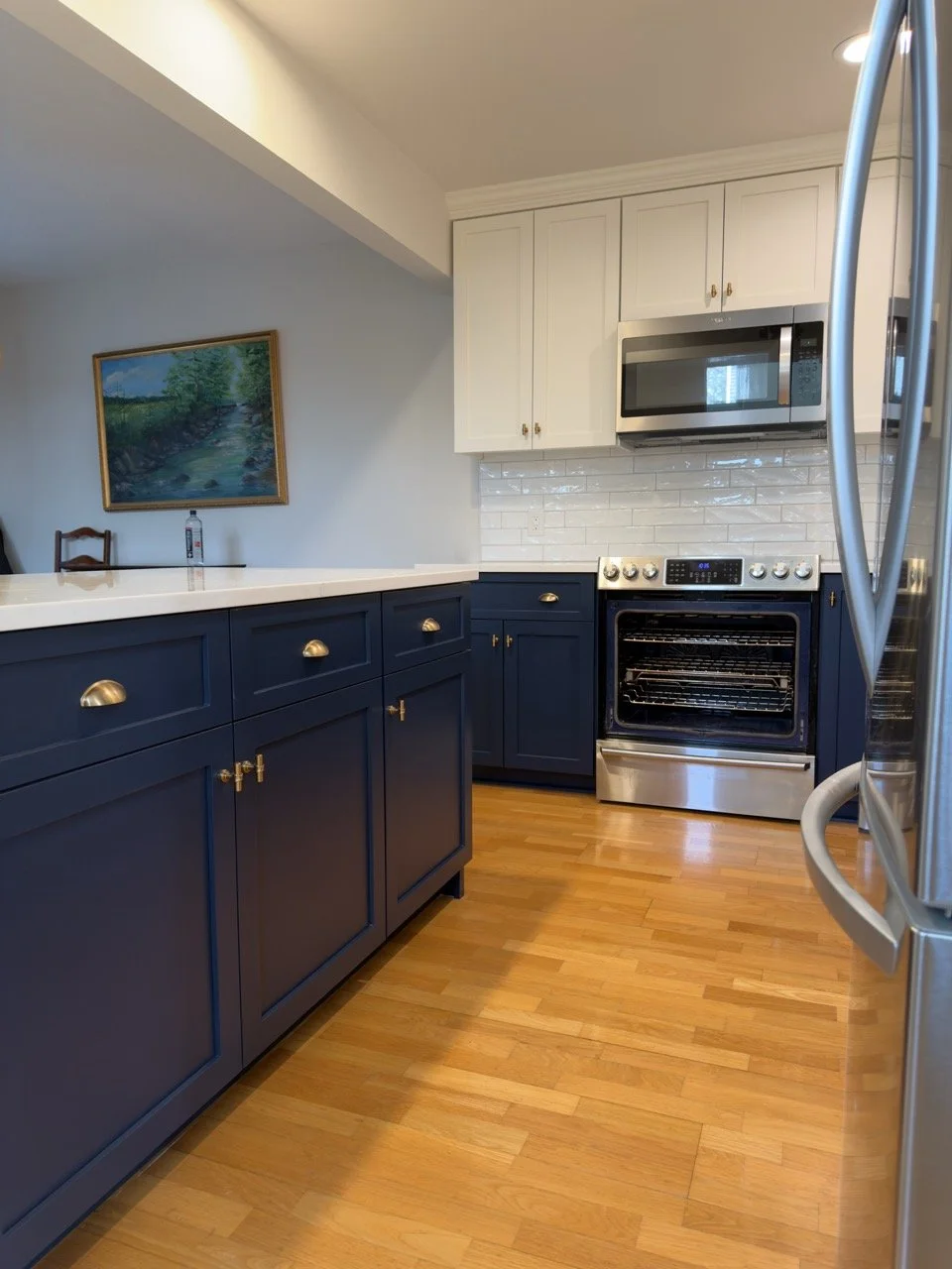 Kitchen with blue lower cabinets and white upper cabinets, stainless steel stove and microwave, white subway tile backsplash, wooden floor, and a painting of a river and trees on the wall.