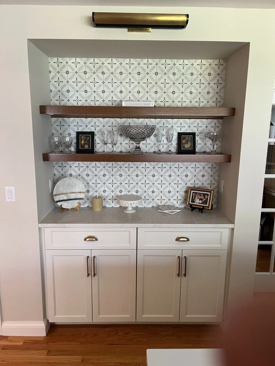 Built-in display cabinet with three open shelves holding decorative glassware, picture frames, and a bowl, against a patterned tile backsplash, with a white cabinet with brass handles below.
