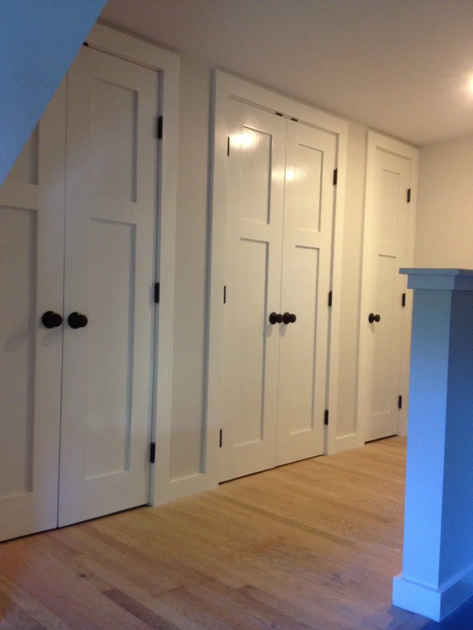 Four white closet doors with black knobs in a room with light-colored wooden flooring and a white wall.