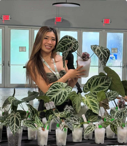Young woman smiling and holding a potted plant with large green leaves, standing behind a table filled with similar plants in small white pots, inside a bright indoor space with glass doors and red exit signs.