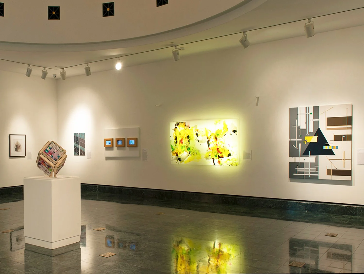 An art gallery with several colorful paintings and artworks displayed on white walls and a white pedestal in the center, featuring a variety of styles including abstract and digital art.