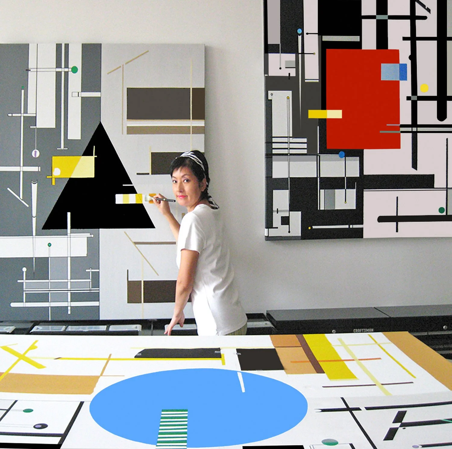 A woman in a white shirt is painting an abstract geometric artwork on a wall with black, white, gray, yellow, and green shapes and lines, in a brightly lit art studio.