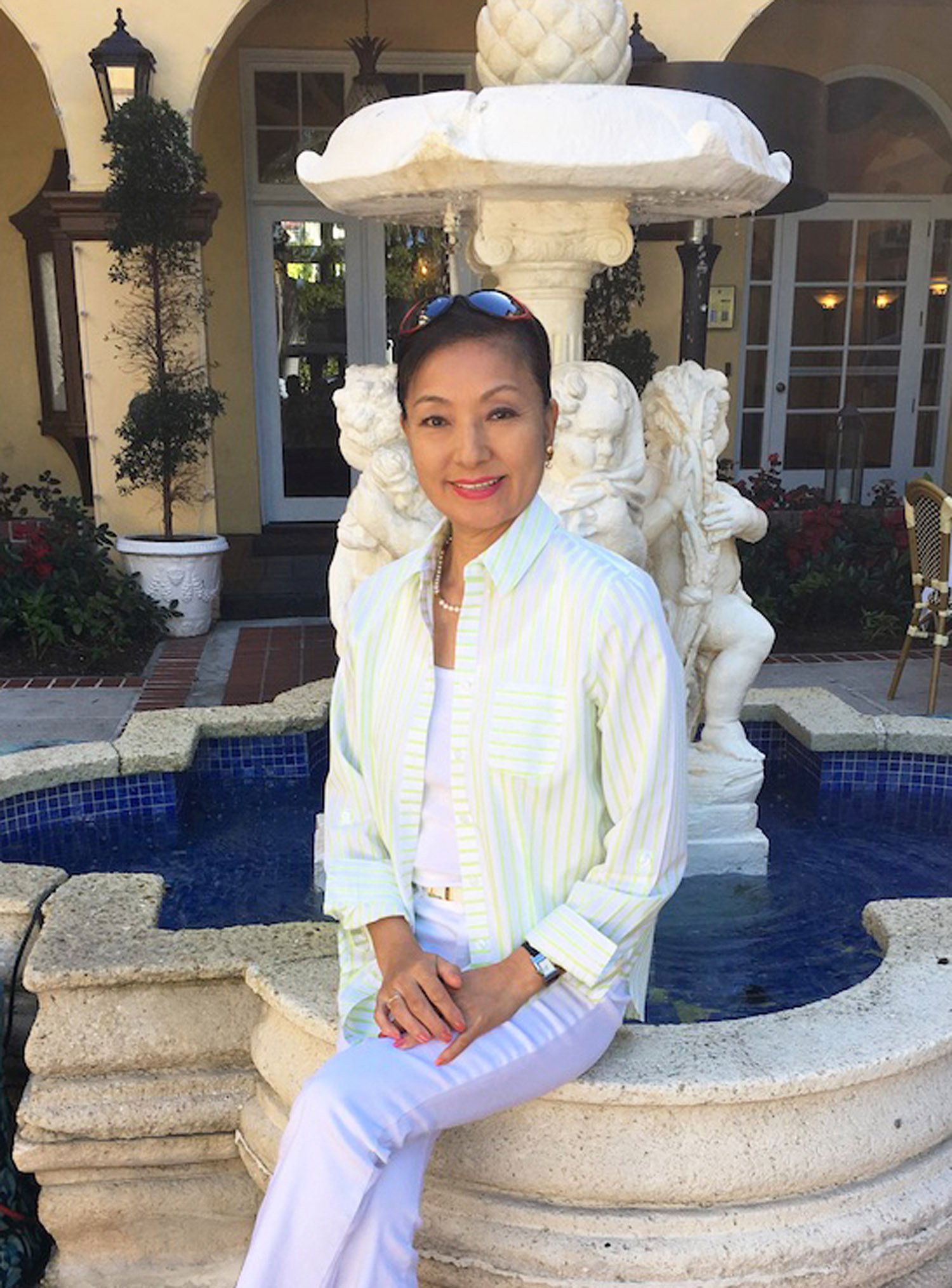 A woman in a yellow and white striped shirt and white pants sitting in front of a large ornate fountain with lion sculptures in an outdoor courtyard.