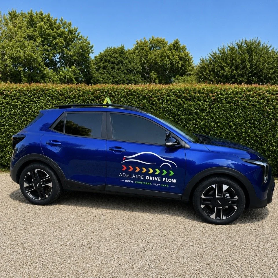 Blue electric vehicle parked on gravel with a logo and text for Adelaide Drive Flow on the side, green trees and bushes in the background, and a clear blue sky.