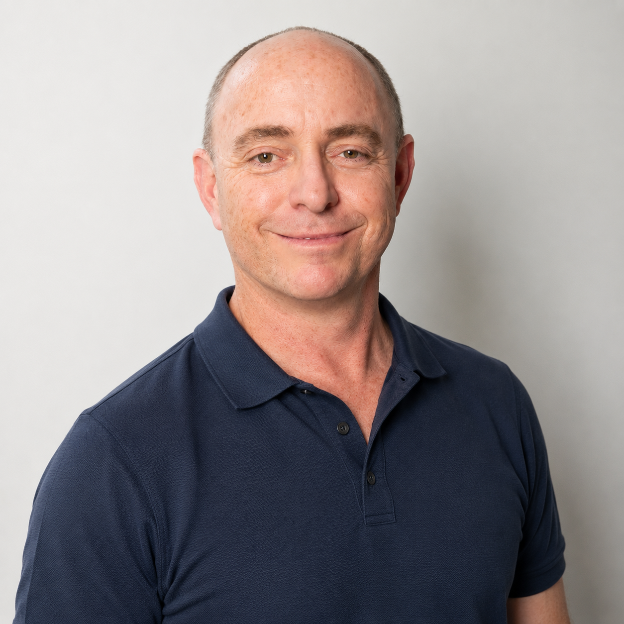 Portrait of a middle-aged man wearing a navy polo shirt, smiling, standing in front of a plain white background.