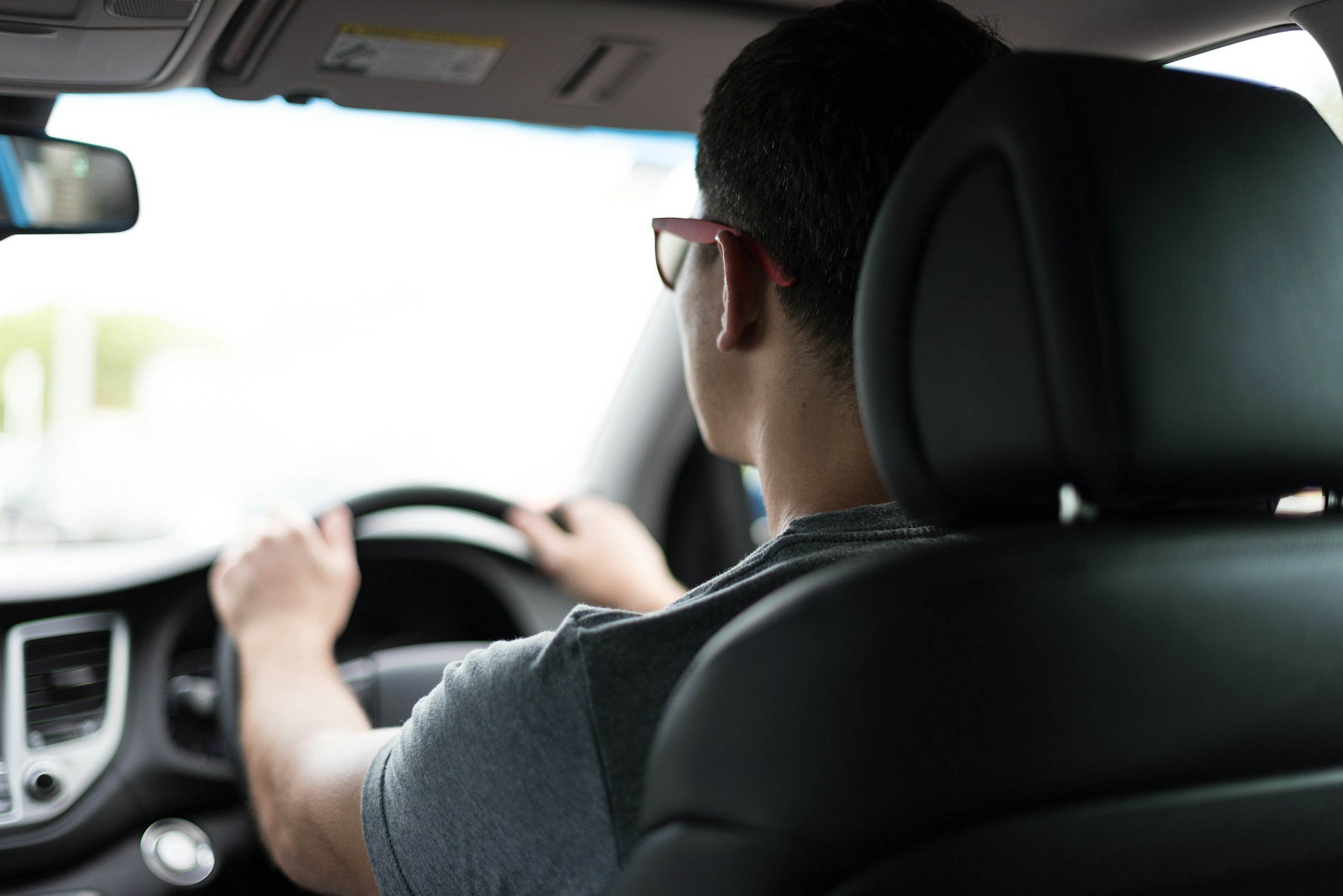Back view of a man driving a car, wearing glasses, with short dark hair, seated in the driver's seat with his hands on the steering wheel.