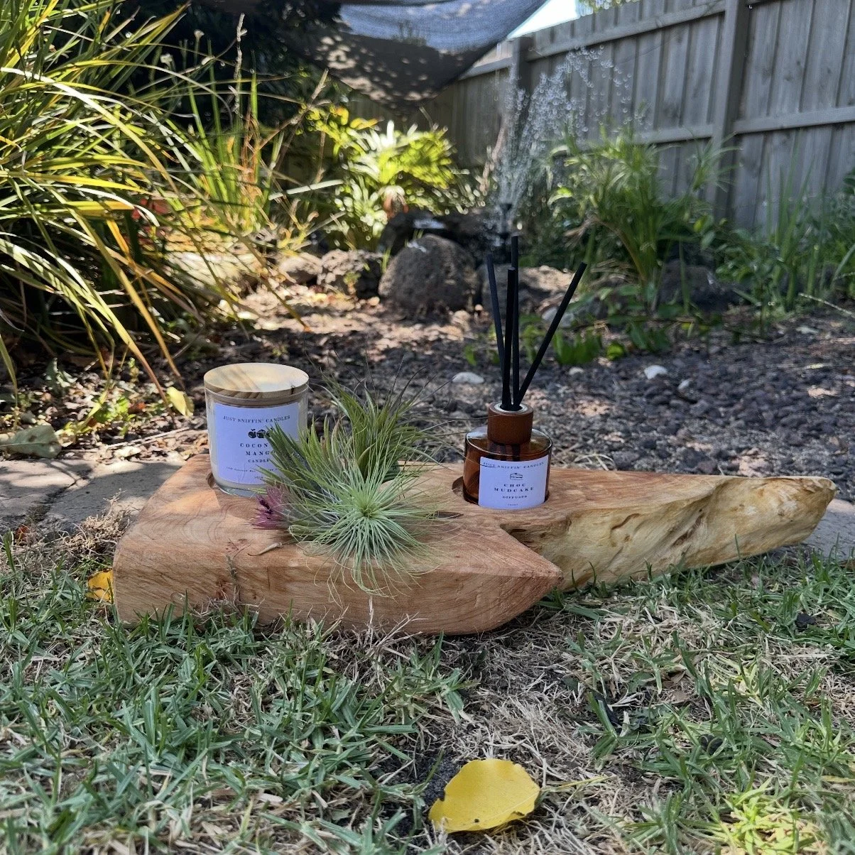 A wooden tray with a cushion on it, placed on grass in a garden. The tray holds a reed diffuser, a candle, and a small plant with purple and green leaves. In the background, there are various plants, rocks, and a wooden fence.