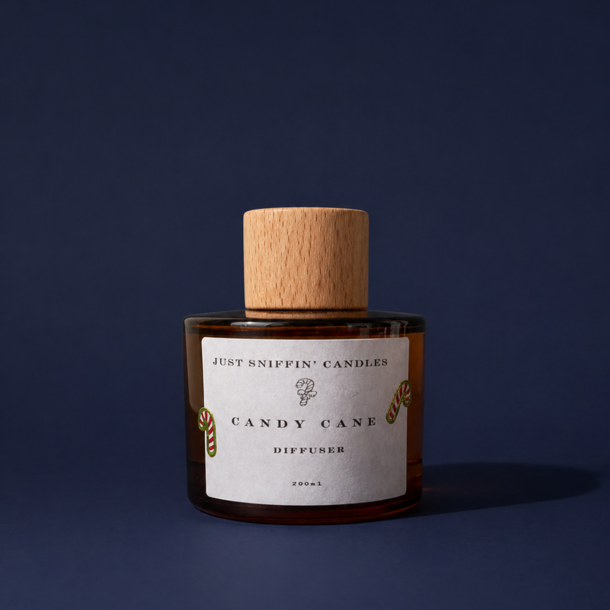 A candle in a brown glass jar with a wooden lid, labeled 'Just Sniffin' Candles Candy Cane Diffuser' with holiday-themed candy cane illustrations, set against a dark blue background.
