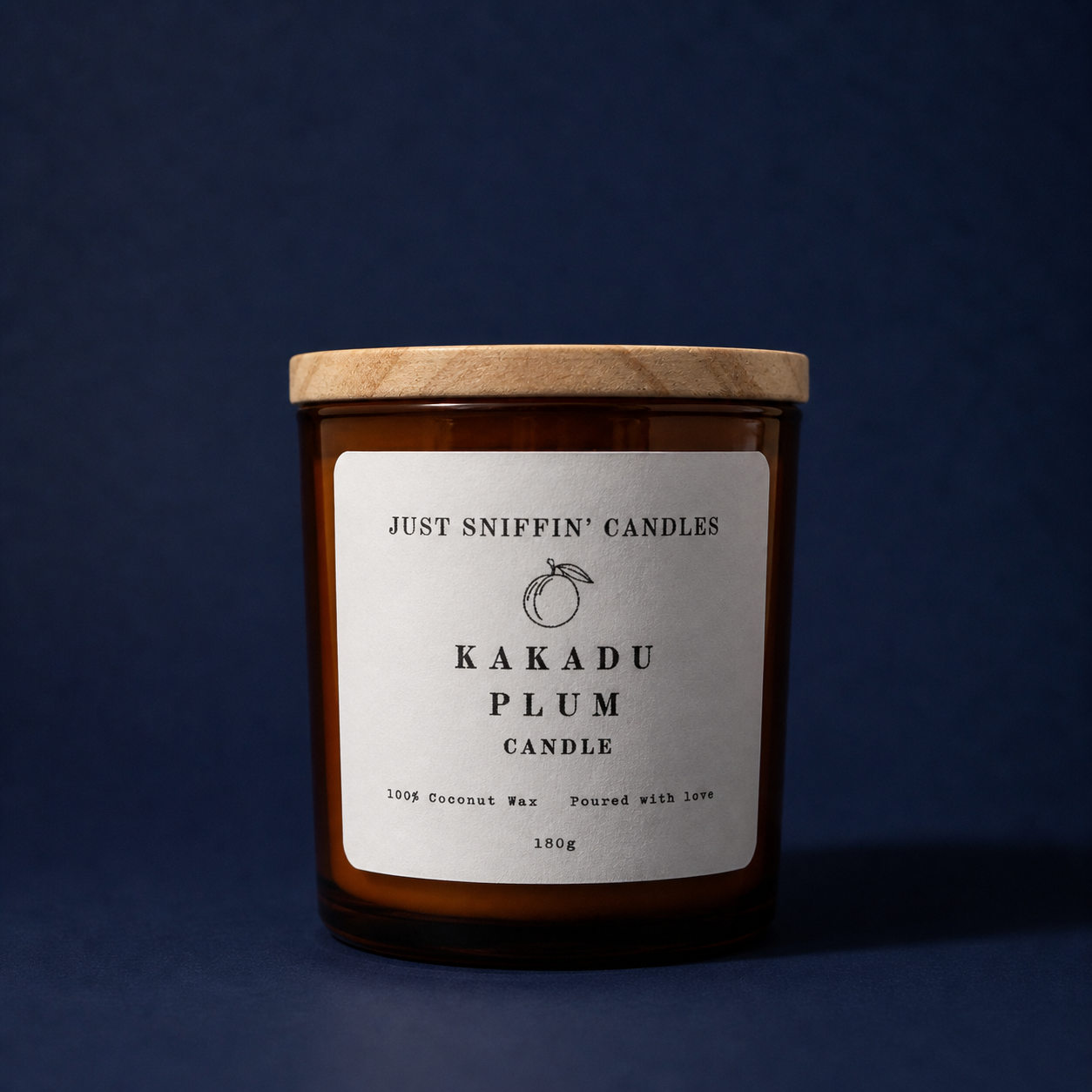 A brown candle jar with a wooden lid, labeled 'Just Sniffin' Candles Kakadu Plum Candle,' made from 100% coconut wax, weighing 180 grams, against a dark blue background.