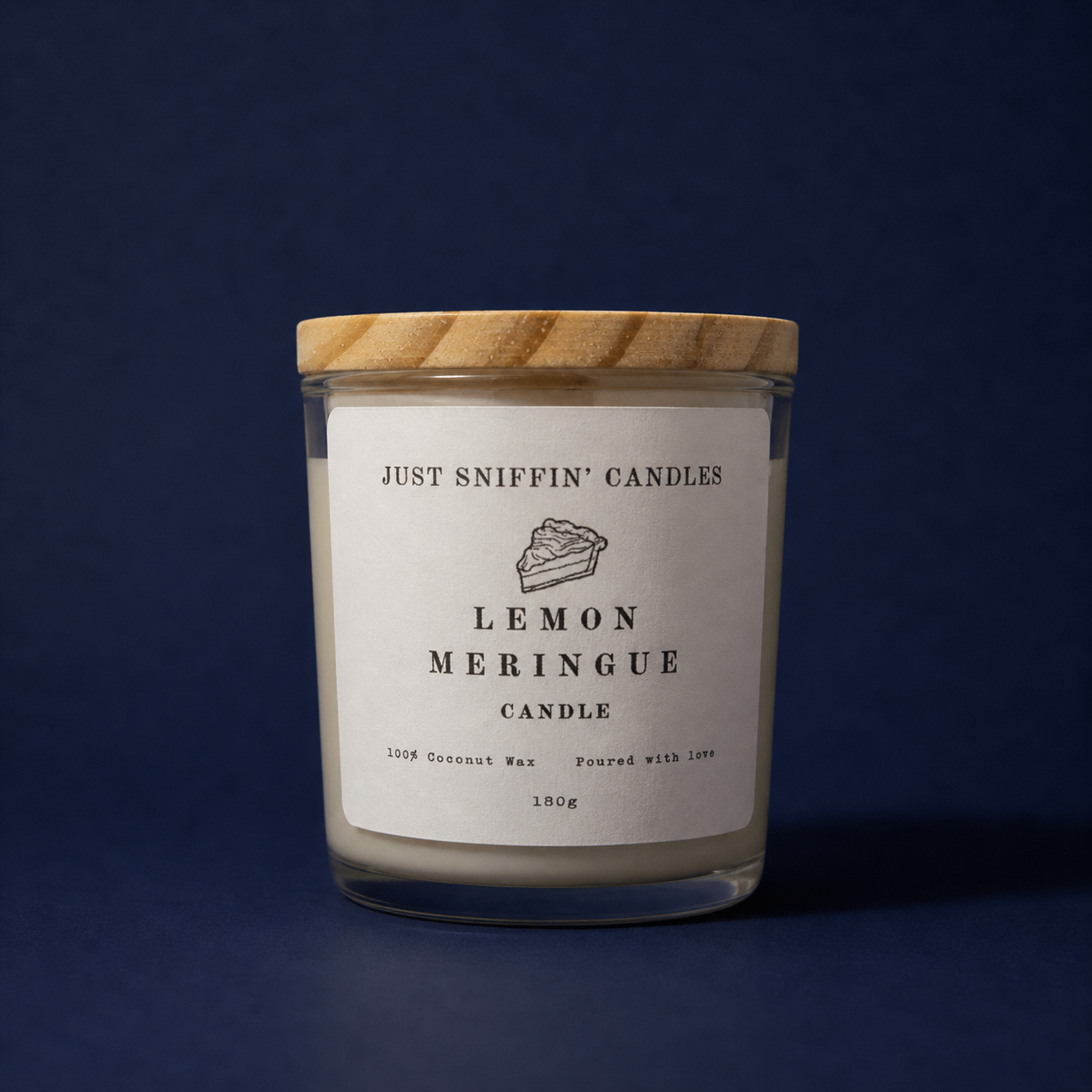 A candle in a clear glass jar with a wooden lid labeled 'Just Sniffin' Candles Lemon Meringue Candle,' made of coconut wax, weighing 180 grams, against a dark blue background.