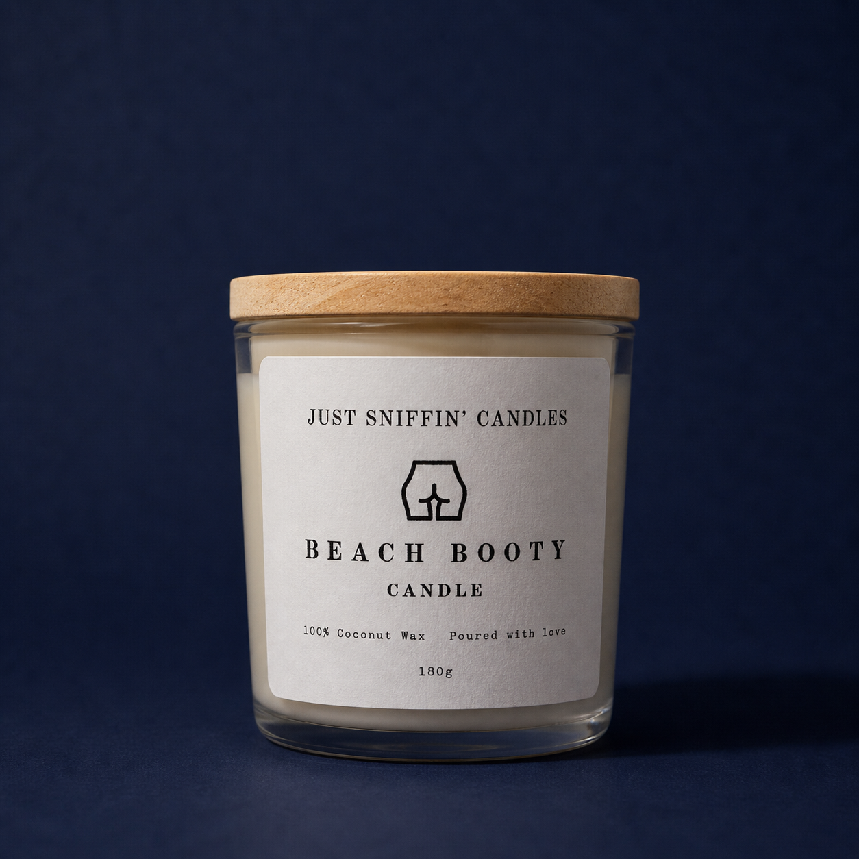 A candle in a glass jar with a wooden lid, labeled "Beach Booty Candle," featuring a minimalist drawing of a woman's hips and buttocks, and text indicating it is made of coconut wax.
