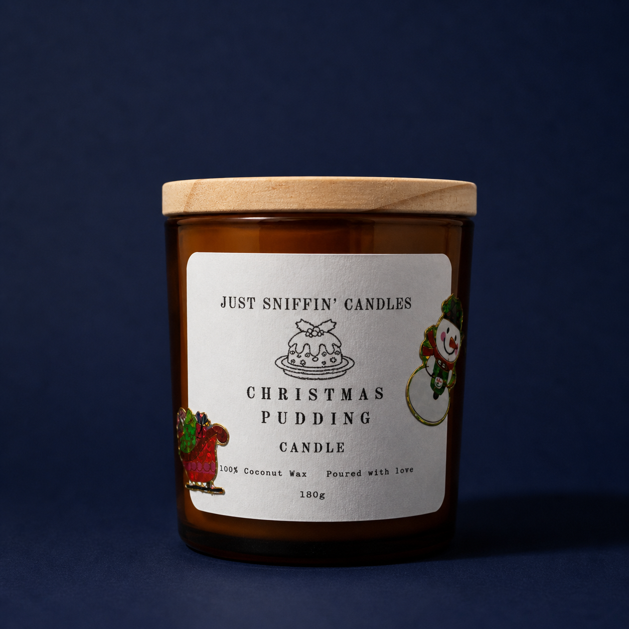 A brown candle jar with a wooden lid labeled "Just Sniffin' Candles Christmas Pudding Candle" featuring Christmas-themed stickers of a snowman and a Santa in a sled, set against a dark blue background.