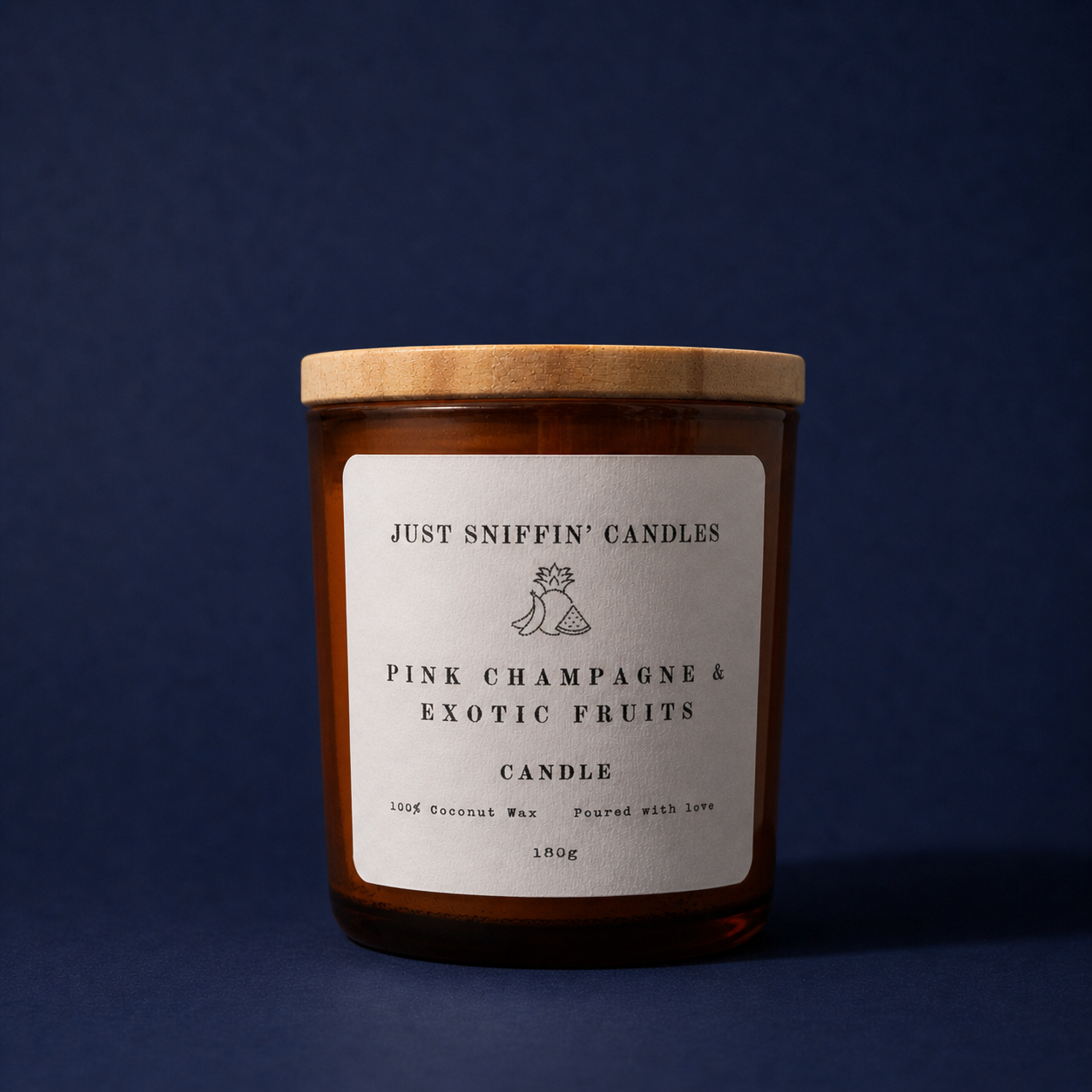 A candle in a brown glass jar with a wooden lid, labeled 'Just Sniffin' Candles' with scent 'Pink Champagne & Exotic Fruits', made from 100% coconut wax, weighing 180 grams, against a dark blue background.