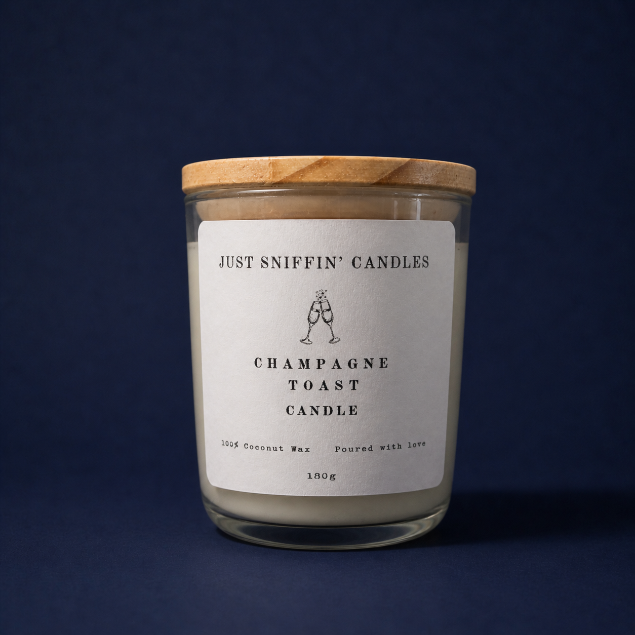 A jar labeled "Just Sniffin' Candles Champagne Toast Candle" with a wooden lid, containing a scented candle made from coconut wax, against a dark blue background.