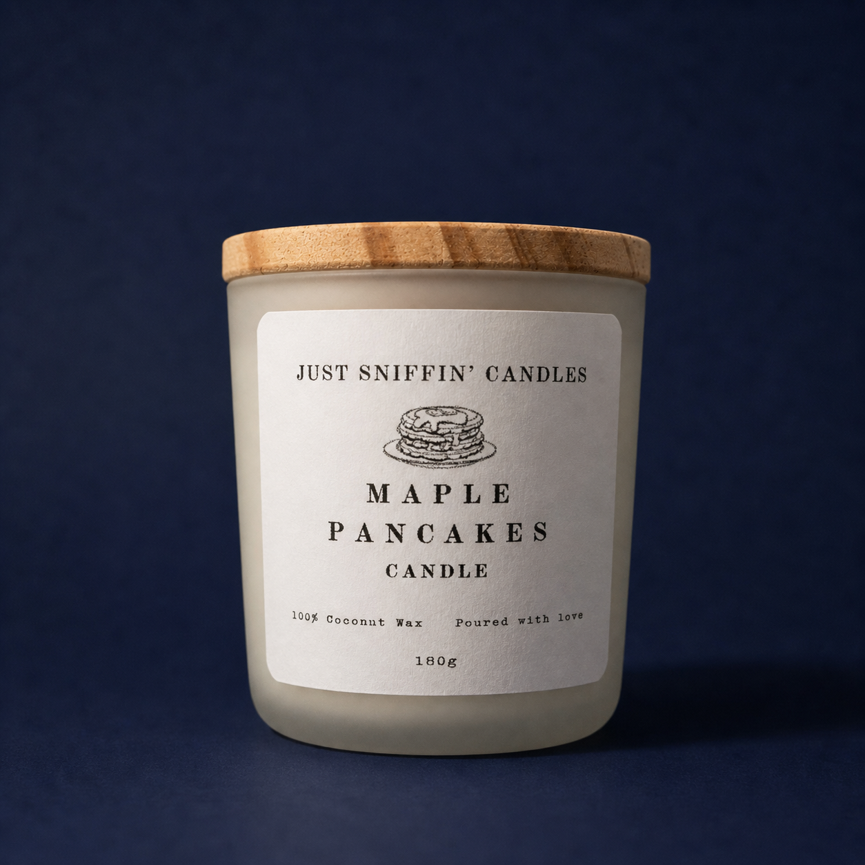 A white candle with a wooden lid, labeled "Just Sniffin' Candles Maple Pancakes Candle," indicating it is made of 100% coconut wax and poured with love, weighing 180 grams, placed against a dark background.