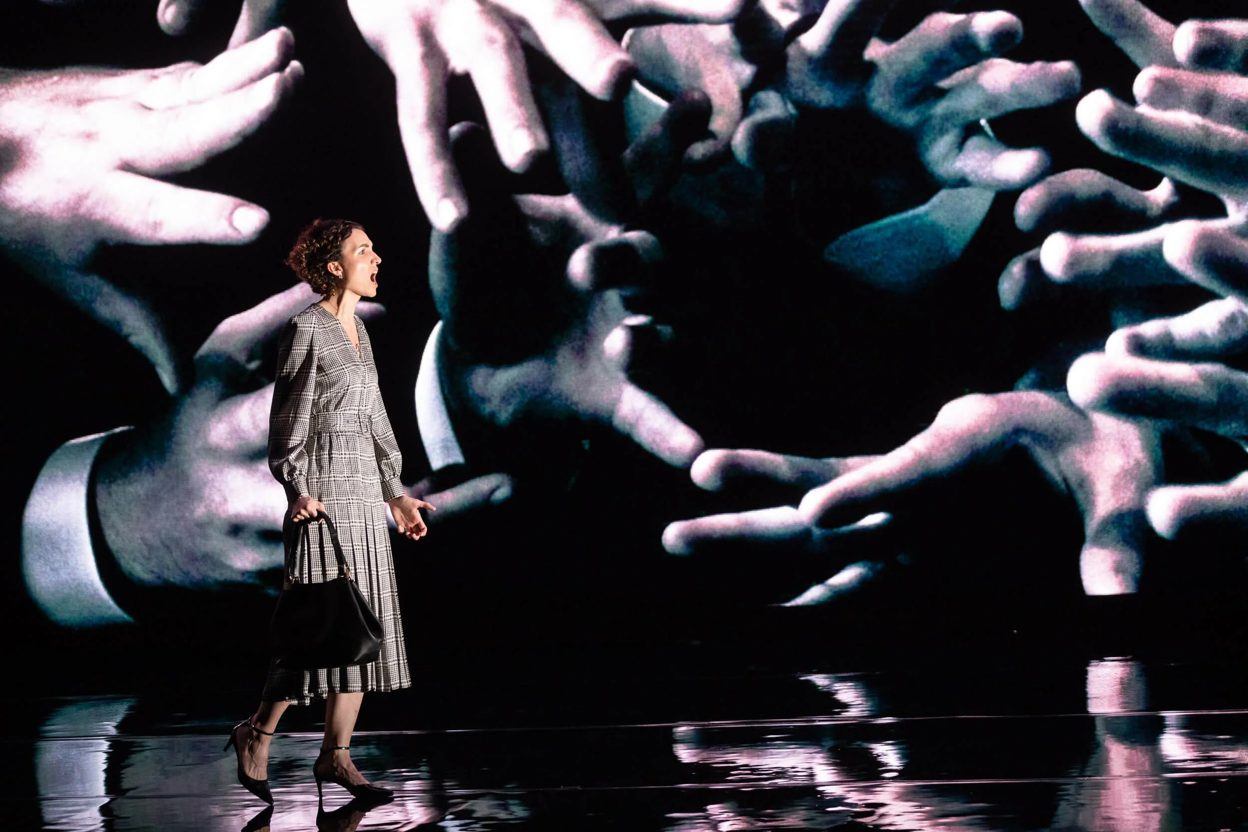 Jessica O'Donoghue with curly hair wearing a plaid dress and high heels is walking on stage with a large screen behind her displaying many hands reaching out in different directions.