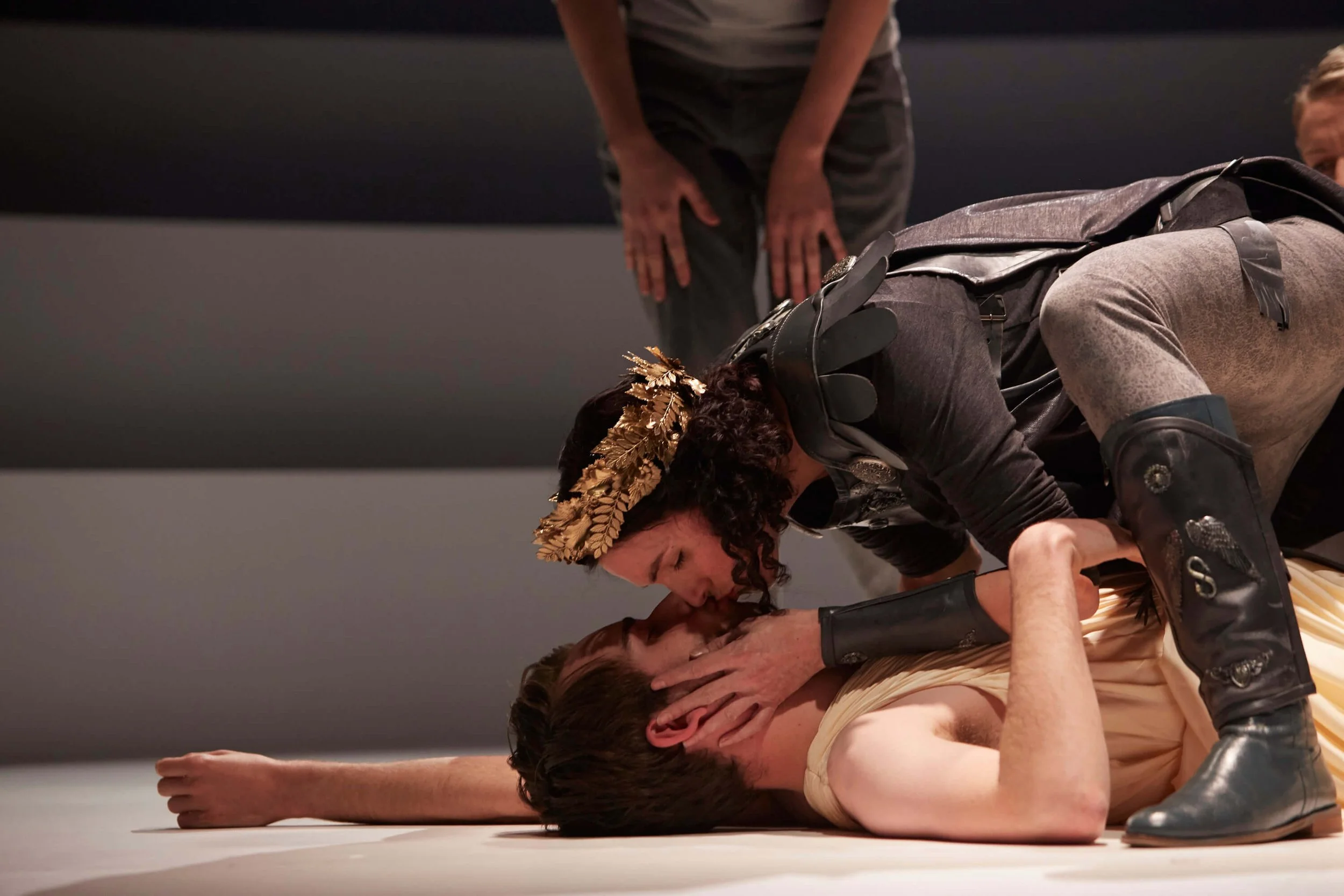 A woman and a man are performing a theatrical scene. The woman, wearing a gold leaf crown and black costume with wings, is leaning over the man, who is lying on the ground. Another person in the background is standing with hands on knees, observing t