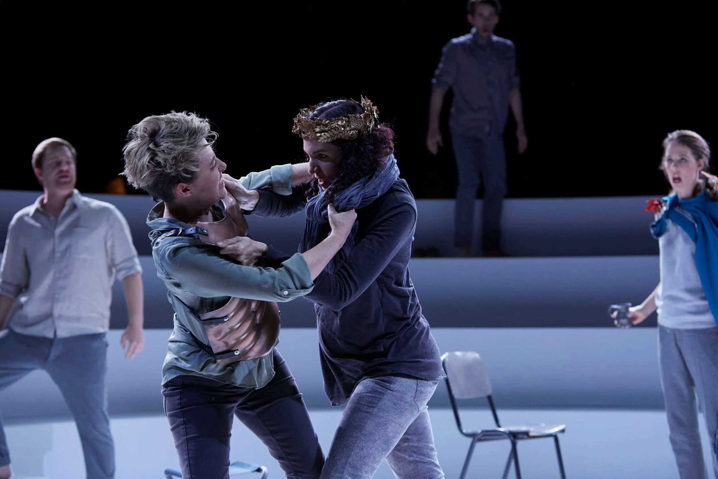 Two women are dancing passionately in a theatrical performance on stage, with three others observing in the background. The scene is set in a dark theater with minimal lighting.