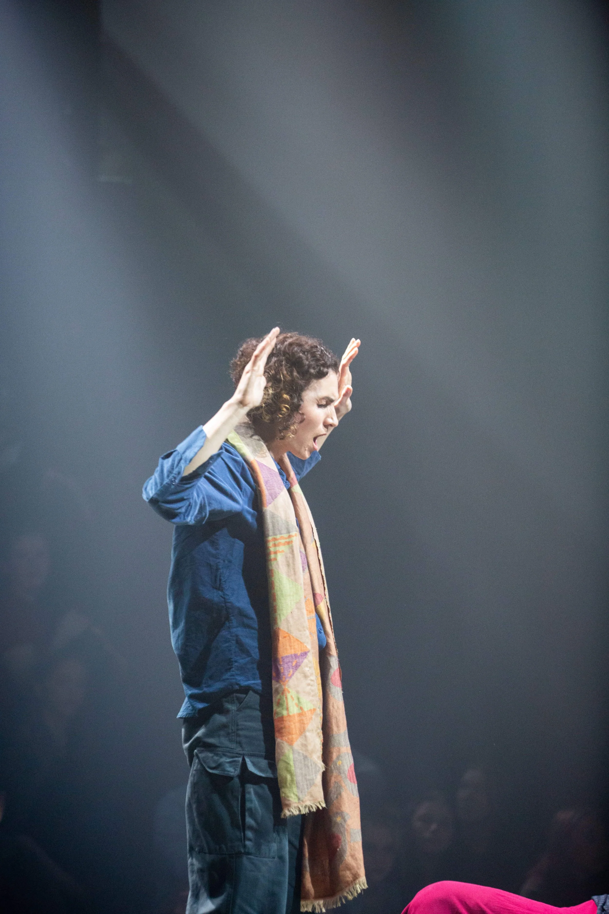 A woman with curly hair is standing with her eyes closed and her hands raised near her head in a gesture of prayer or meditation. She is wearing a blue jacket and a patterned scarf. The background is dark and out of focus.