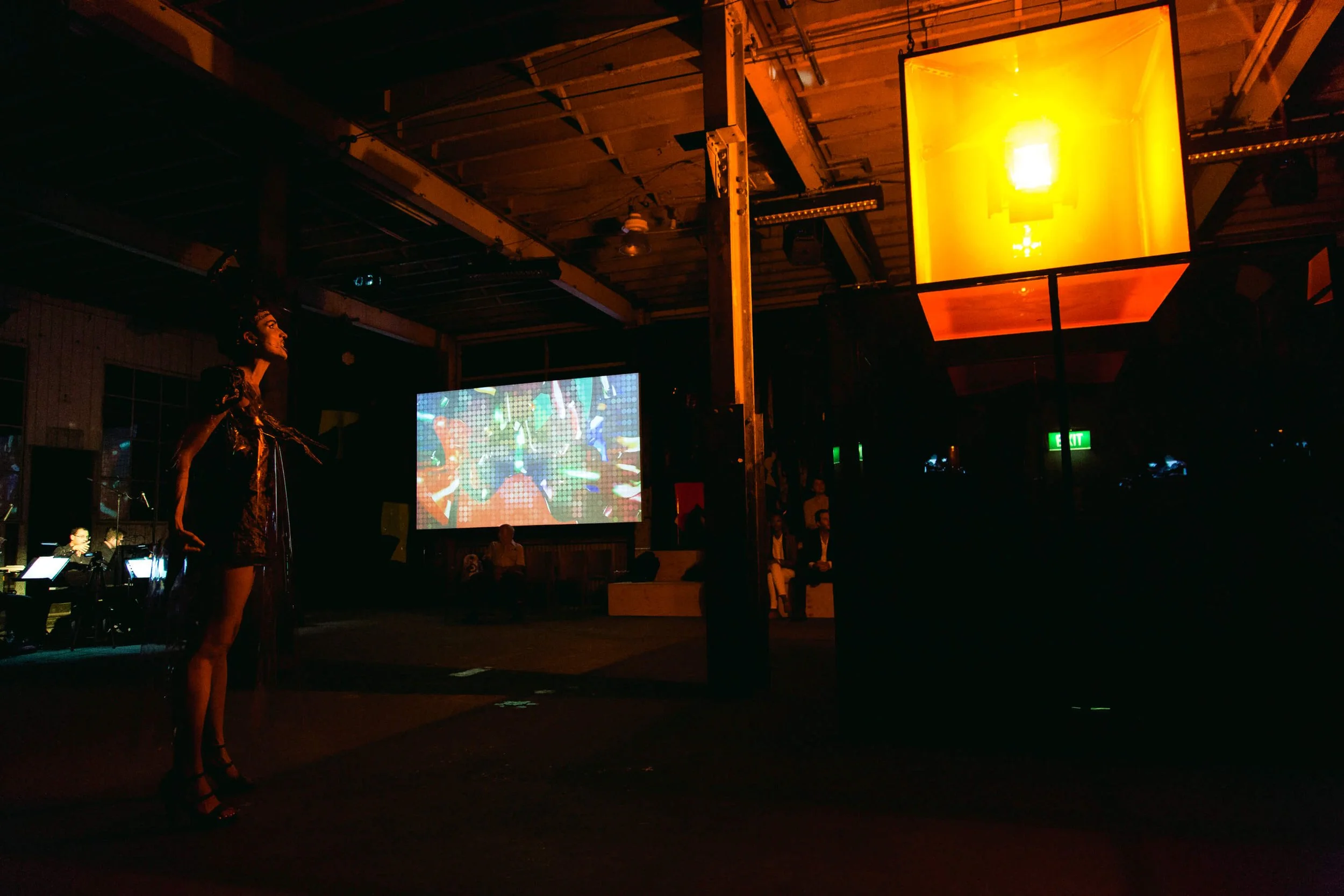 A woman standing on stage with a microphone, in a dimly lit venue with a large screen showing an animated digital display, and an orange hexagonal light fixture overhead.