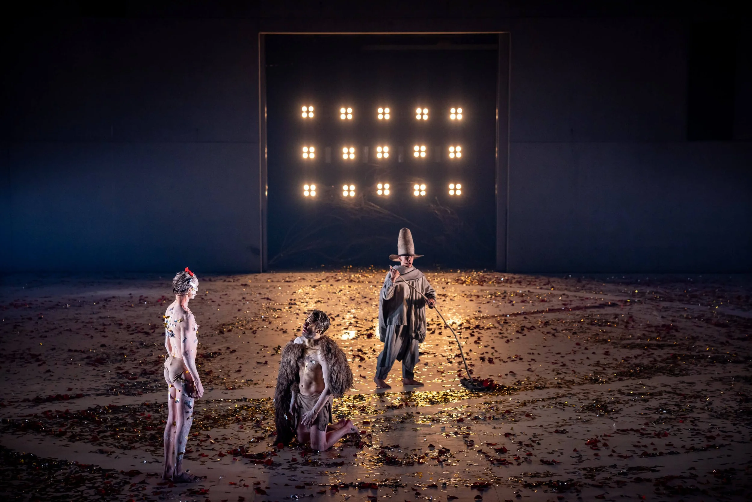 The image depicts a theatrical scene with a dark background. In the scene, there are four models dressed in elaborate costumes that resemble fantastical or mythical creatures, standing or kneeling on a stage scattered with gold and red debris.