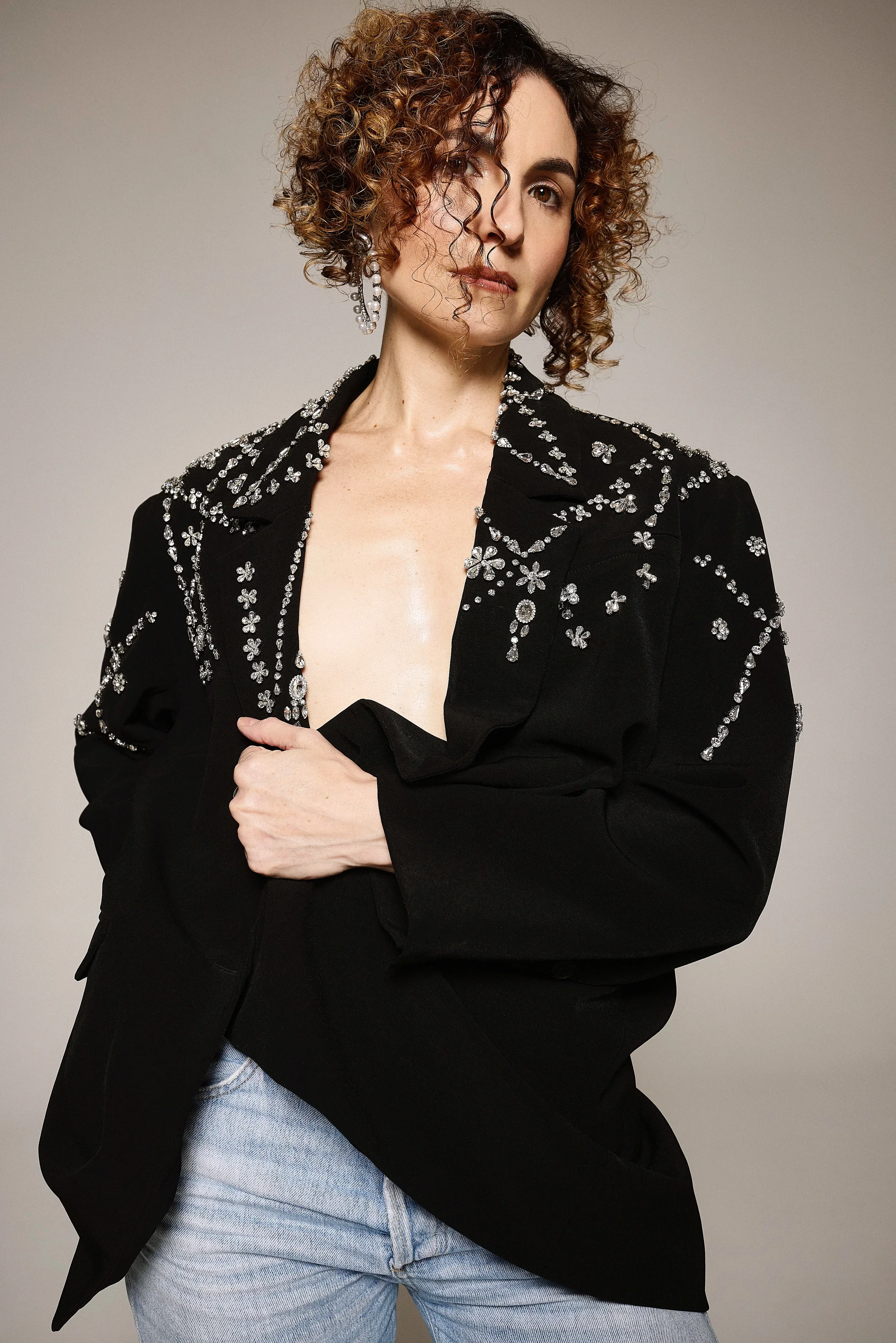 Jessica O'Donoghue with curly brown hair, wearing a black jacket adorned with rhinestones and earrings, poses against a plain background.