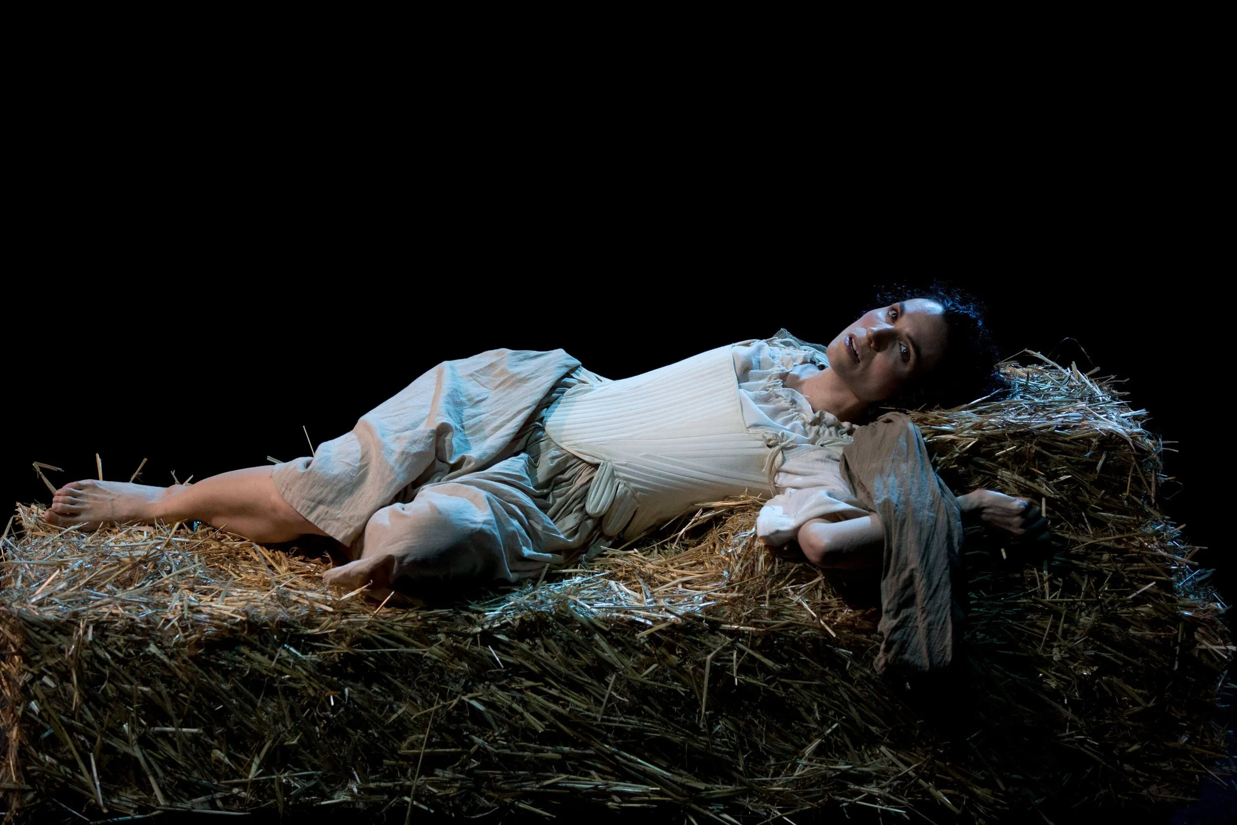 Jessica O'Donoghue lying on her side on a bed of straw, wearing a light-colored dress, with a worried expression, in a dark setting.