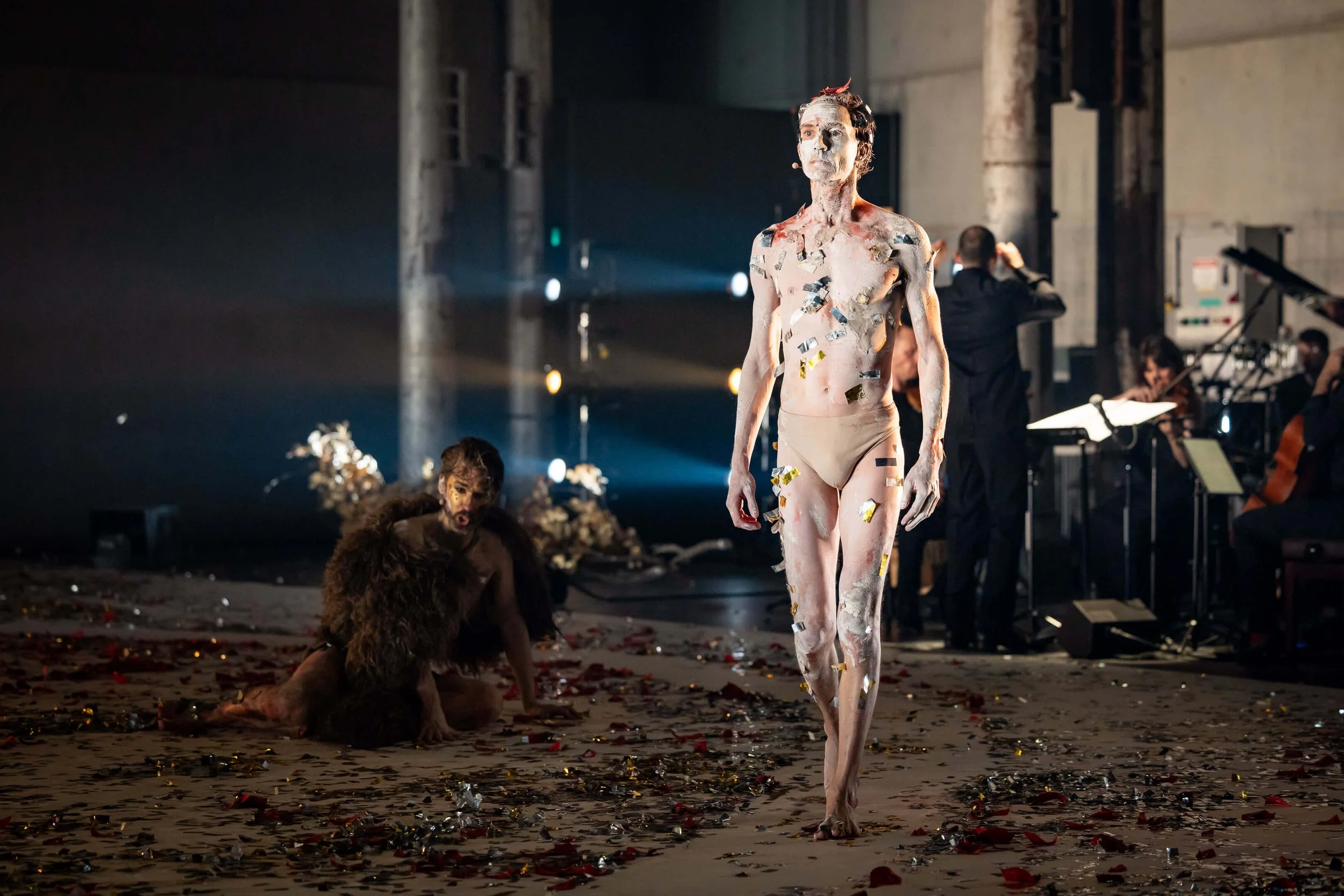 A performer covered in reflective confetti walks across a stage, with others sitting or lying on the floor amidst scattered confetti and debris, in a dimly lit, industrial-style theater.