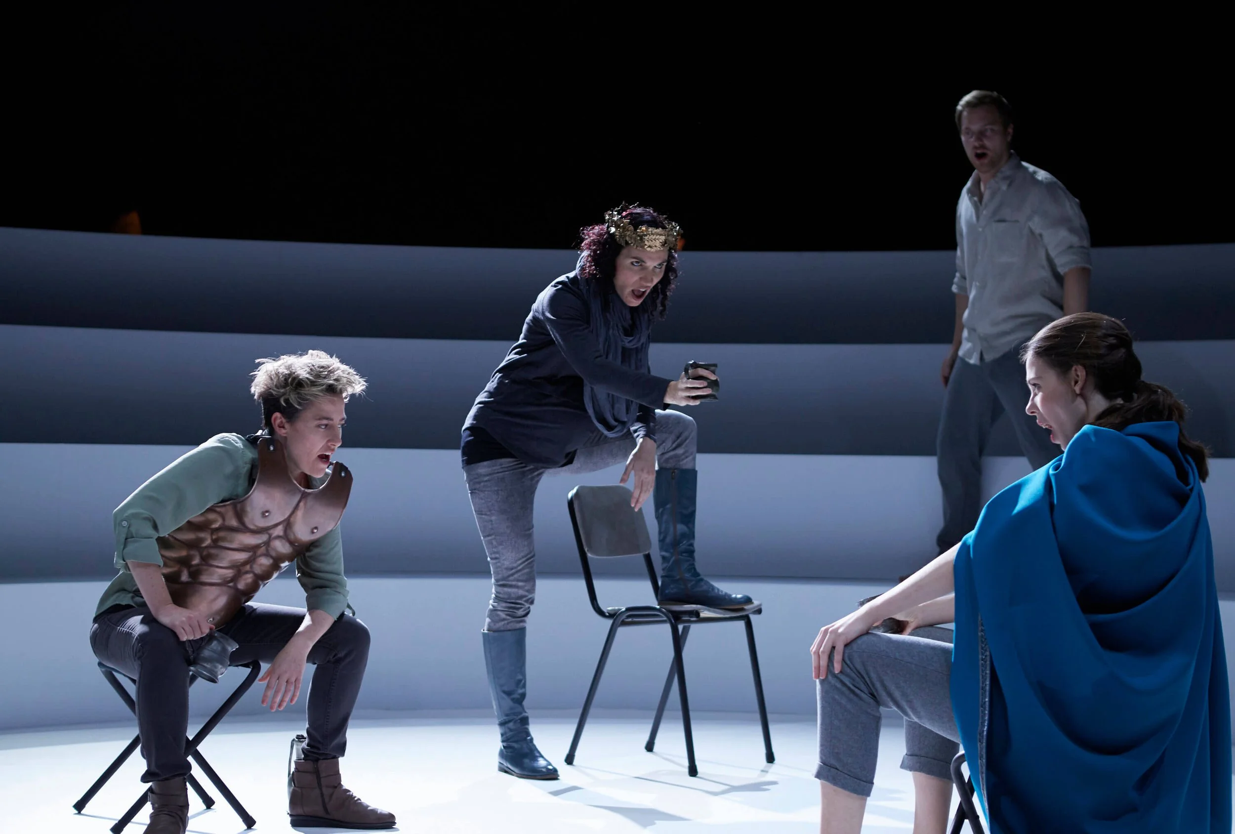 Group of five young adults in dramatic poses on a dark stage, some sitting on chairs, others standing, with intense facial expressions and casual clothing.