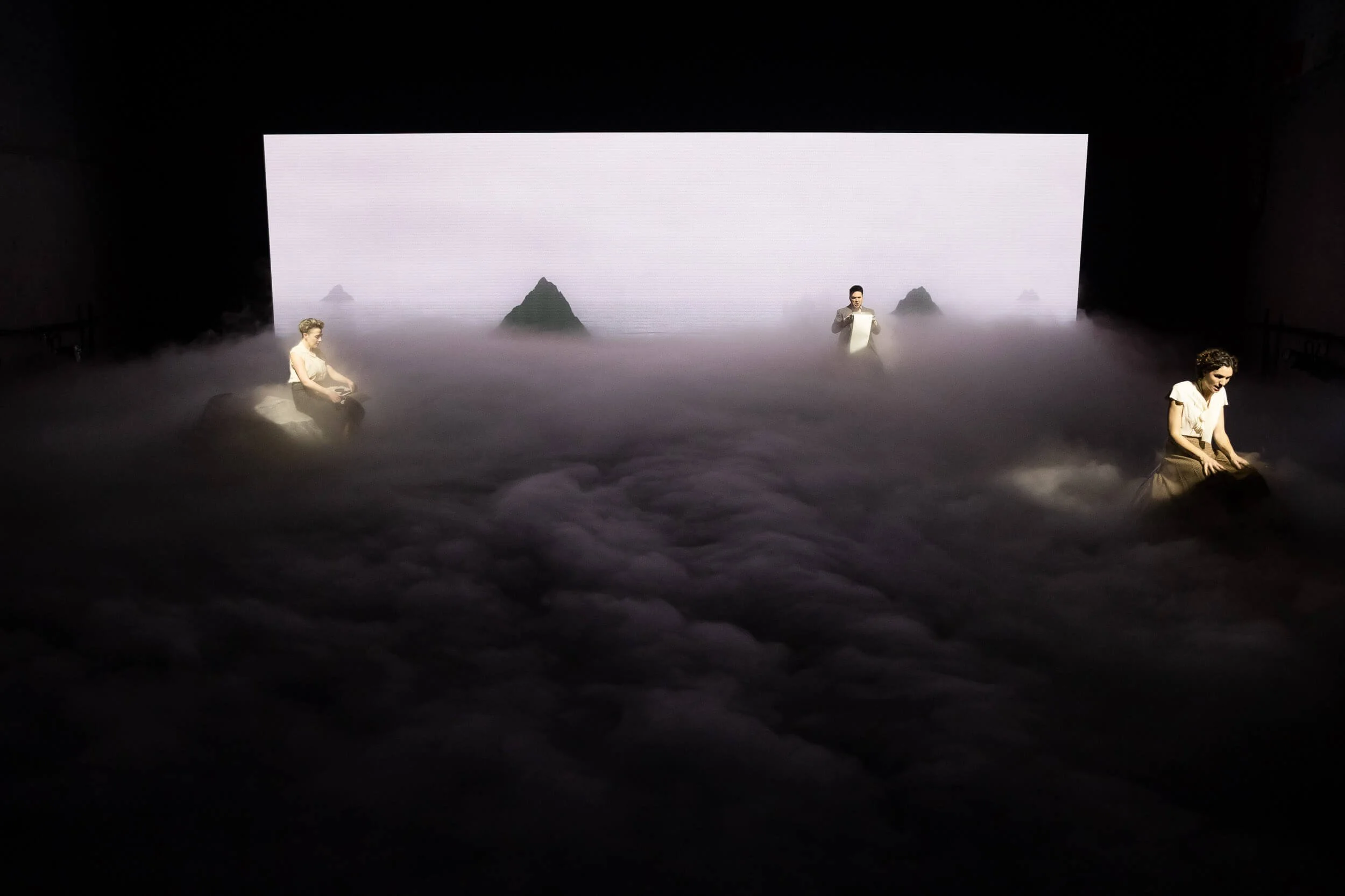 A stage with a large digital screen showing a foggy landscape with distant mountains. Three performers are sitting or standing among fake clouds or fog effects, with one standing and reading from a paper.