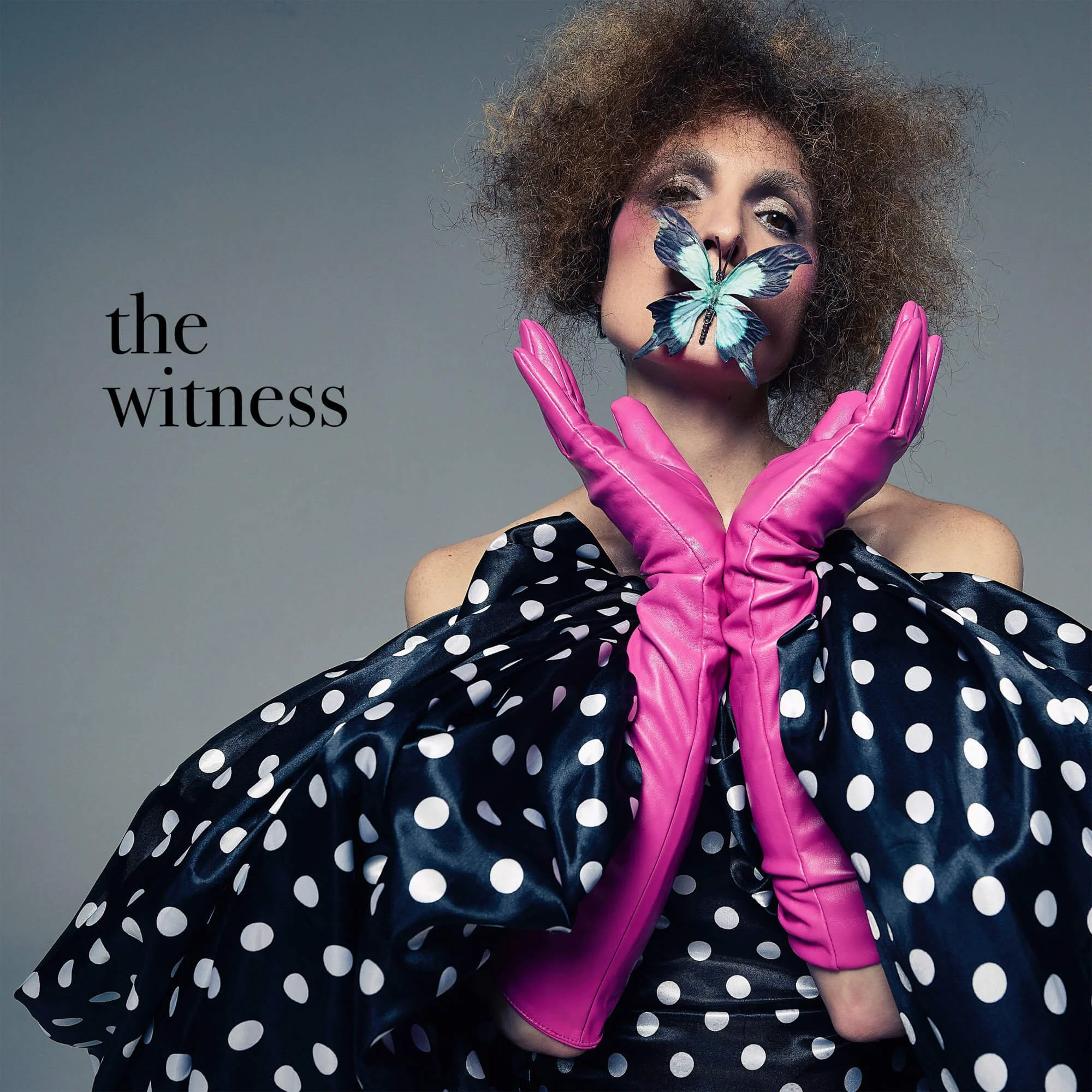 Jessica O'Donoghue with curly hair and makeup covering one eye, wearing bright pink gloves, a polka dot dress, and holding a large blue butterfly on her face. The background is gray.