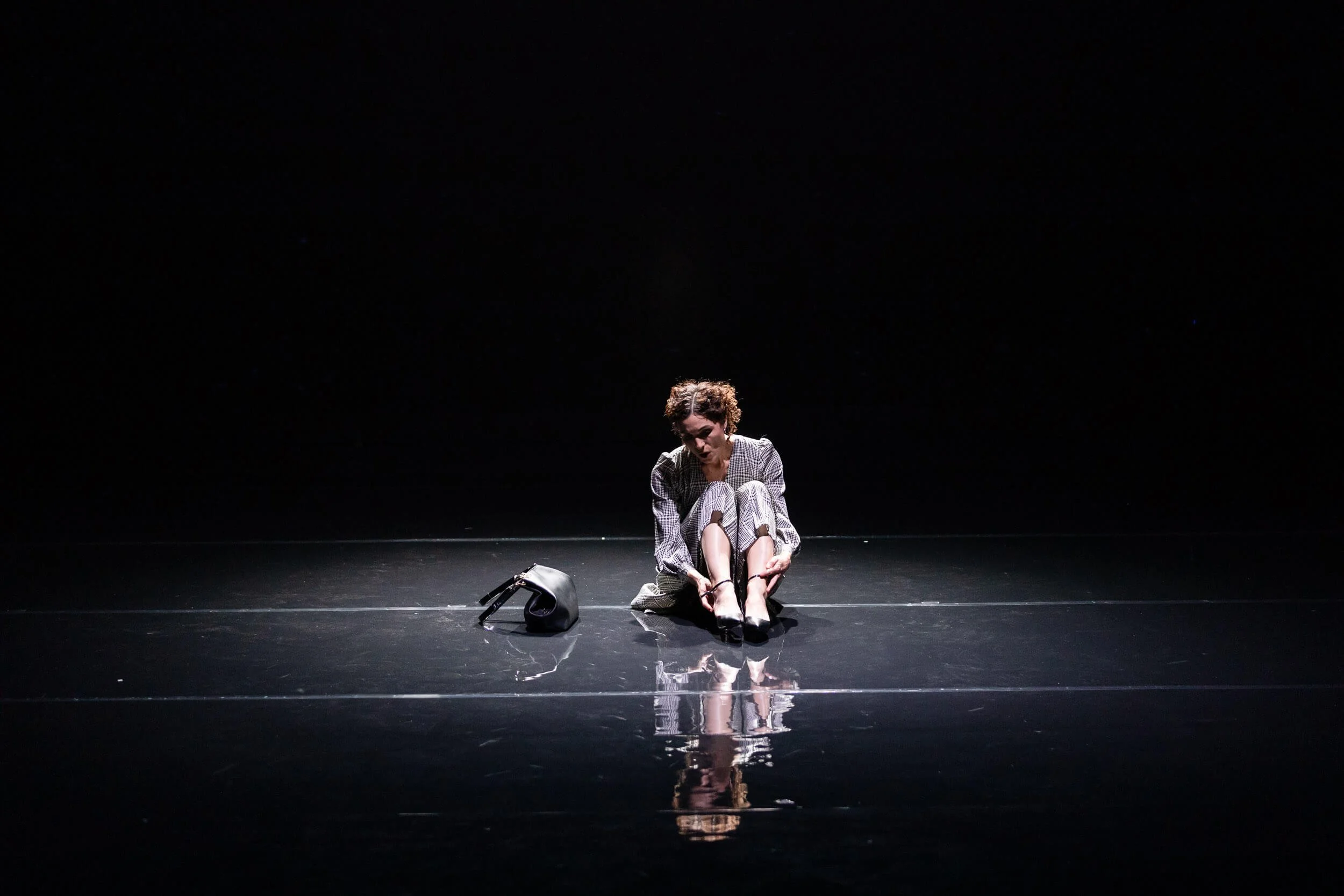 A woman sitting on the stage floor, putting on black high heels. A black handbag is on the floor next to her, and the background is dark.