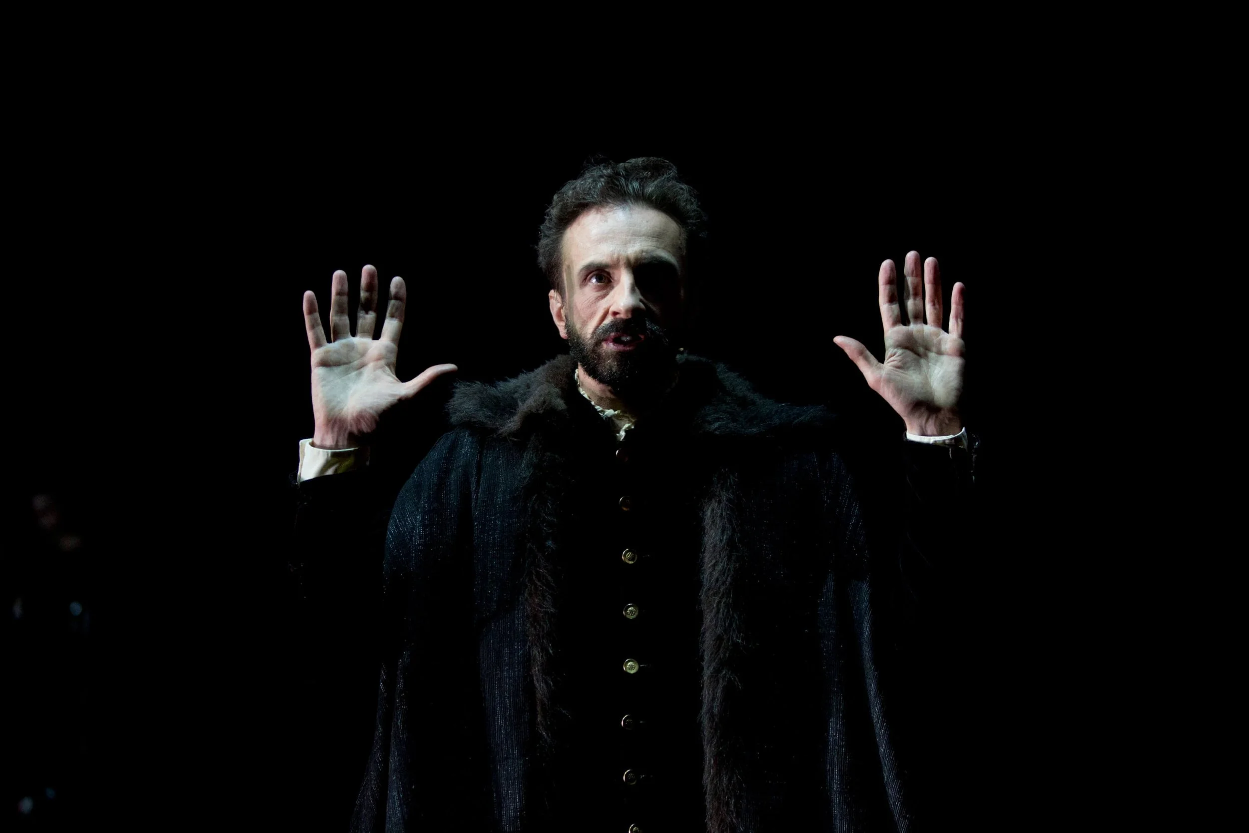 A man with a beard and dark hair, dressed in dark clothing, is raising both hands with his palms open against a dark background.