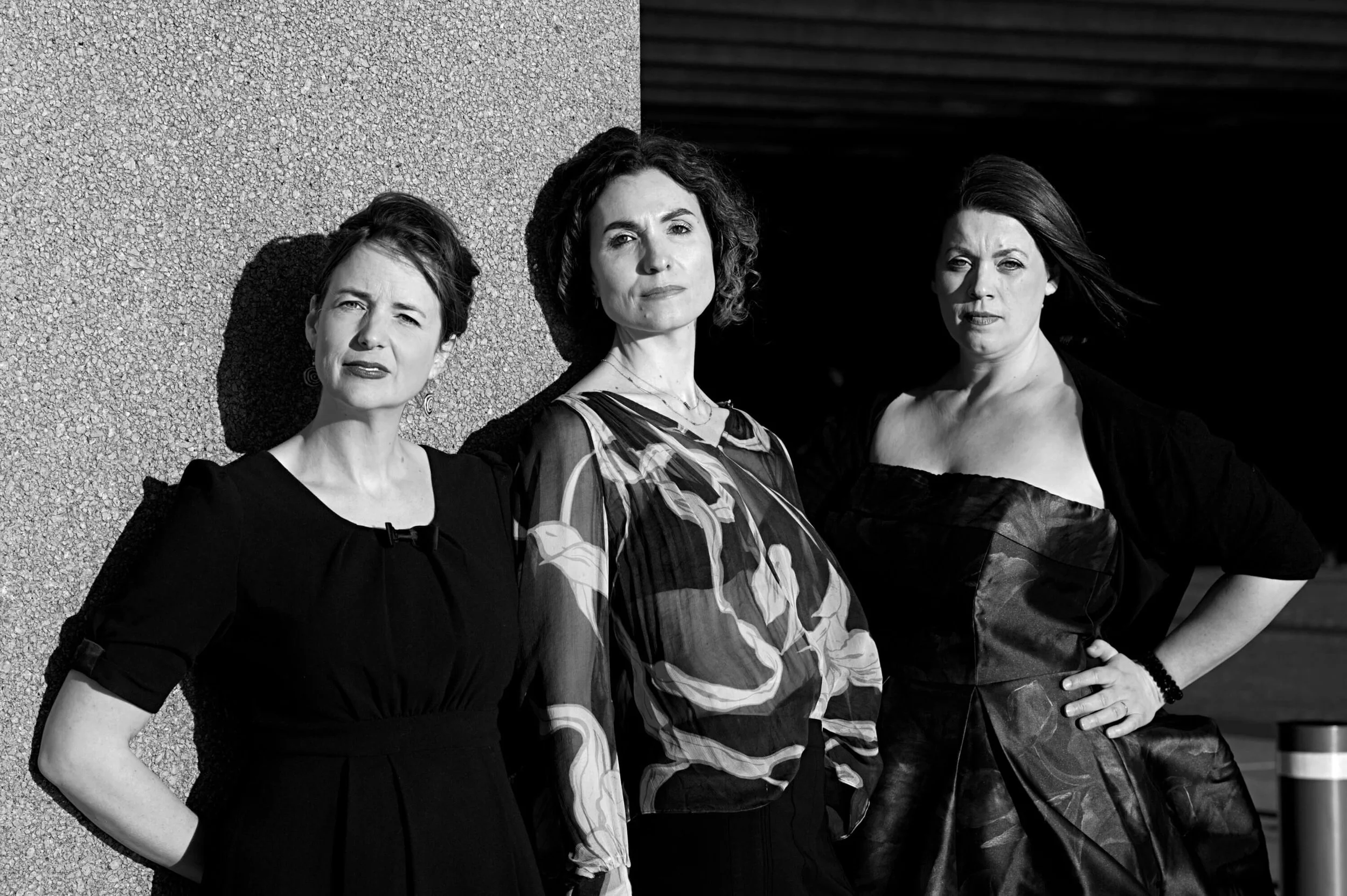 Three women standing outdoors against a textured wall, with sunlight casting shadows on their faces and a dark background on the right.