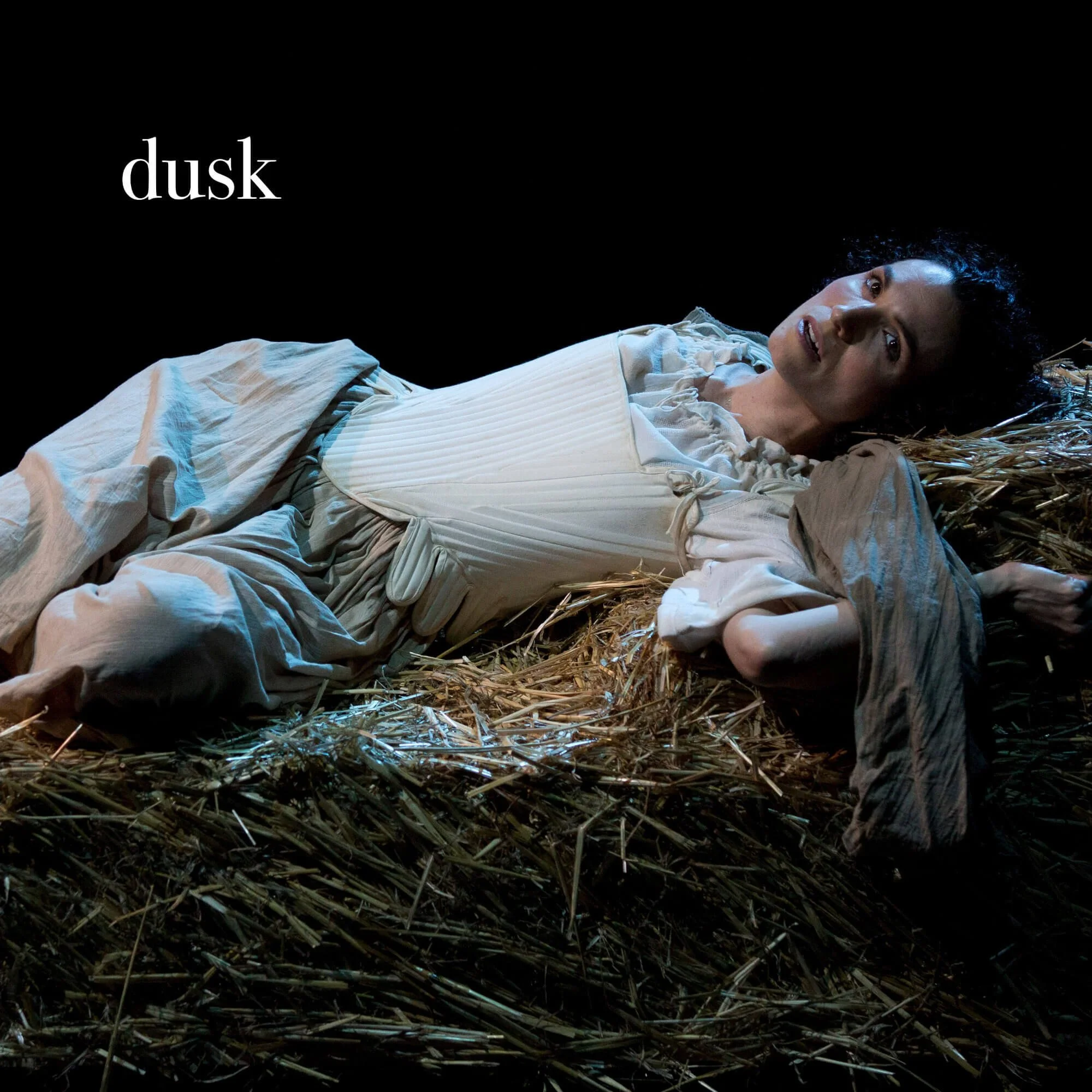 Jessica O'Donoghue lying on a bed of hay in a dark setting, looking up with an expression of surprise or curiosity, wearing a light-colored dress, with the word 'dusk' in white text on a black background.