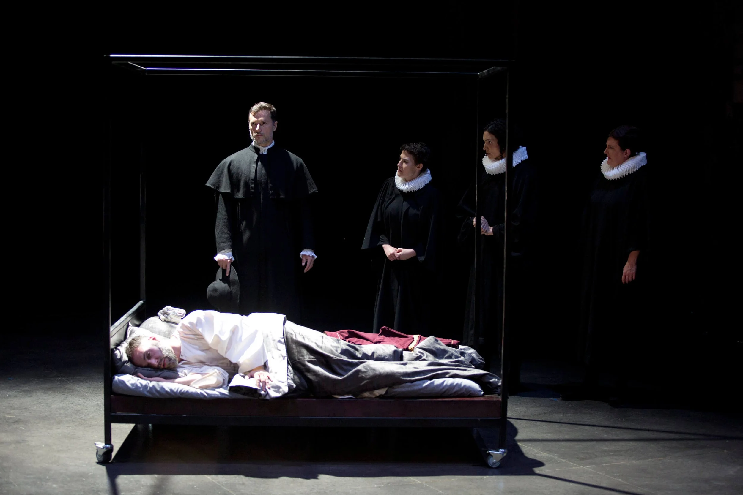 Scene from a theatrical play showing a man lying in a bed with a blanket, surrounded by a mirror reflecting four people in black robes with white ruffled collars, one of whom appears to be a priest, in a dark stage setting.