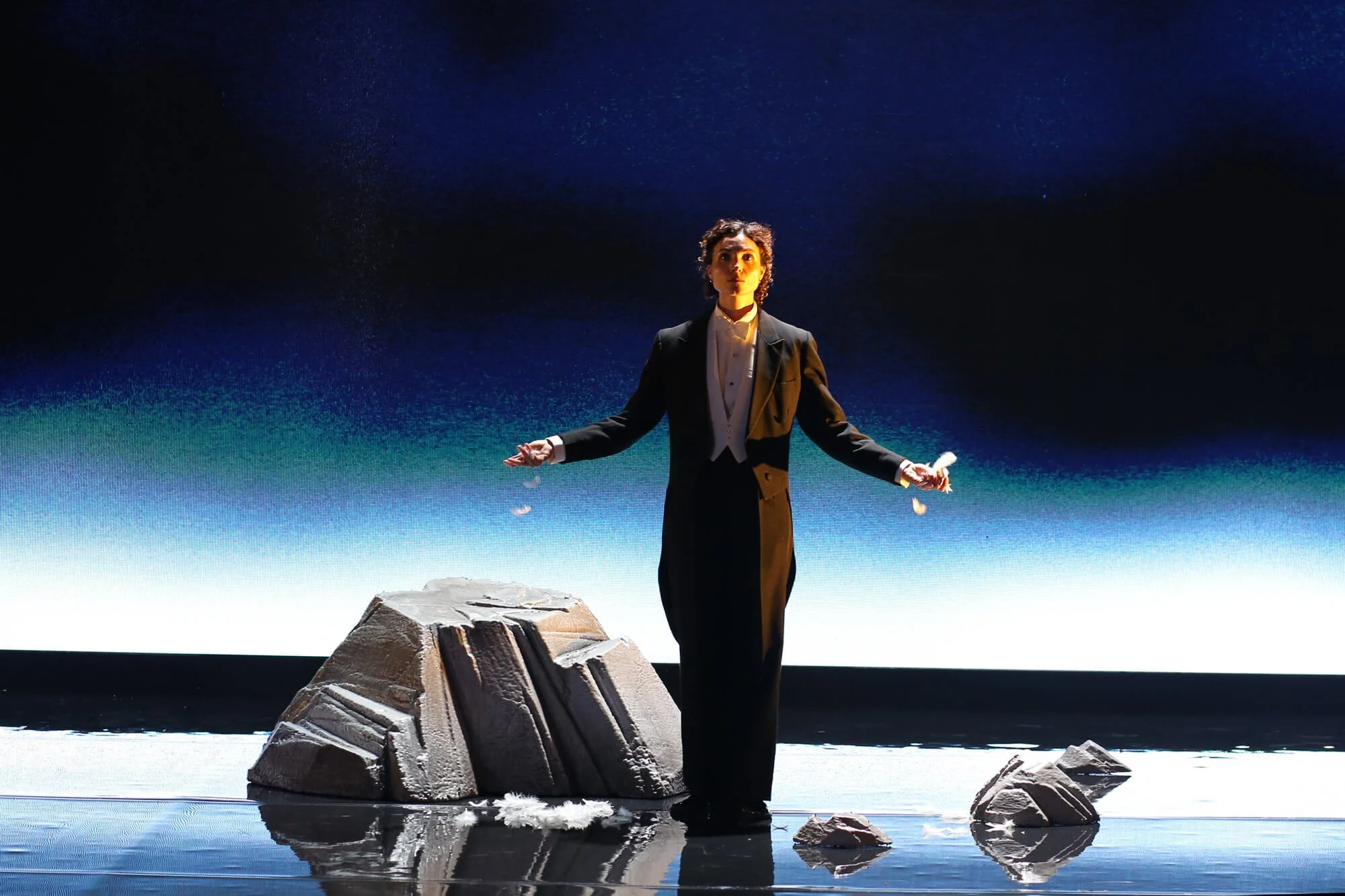 Jessica O'Donoghue in a black tuxedo and white shirt standing on a rocky surface underwater, with a starry night sky visible above and reflections in the water.